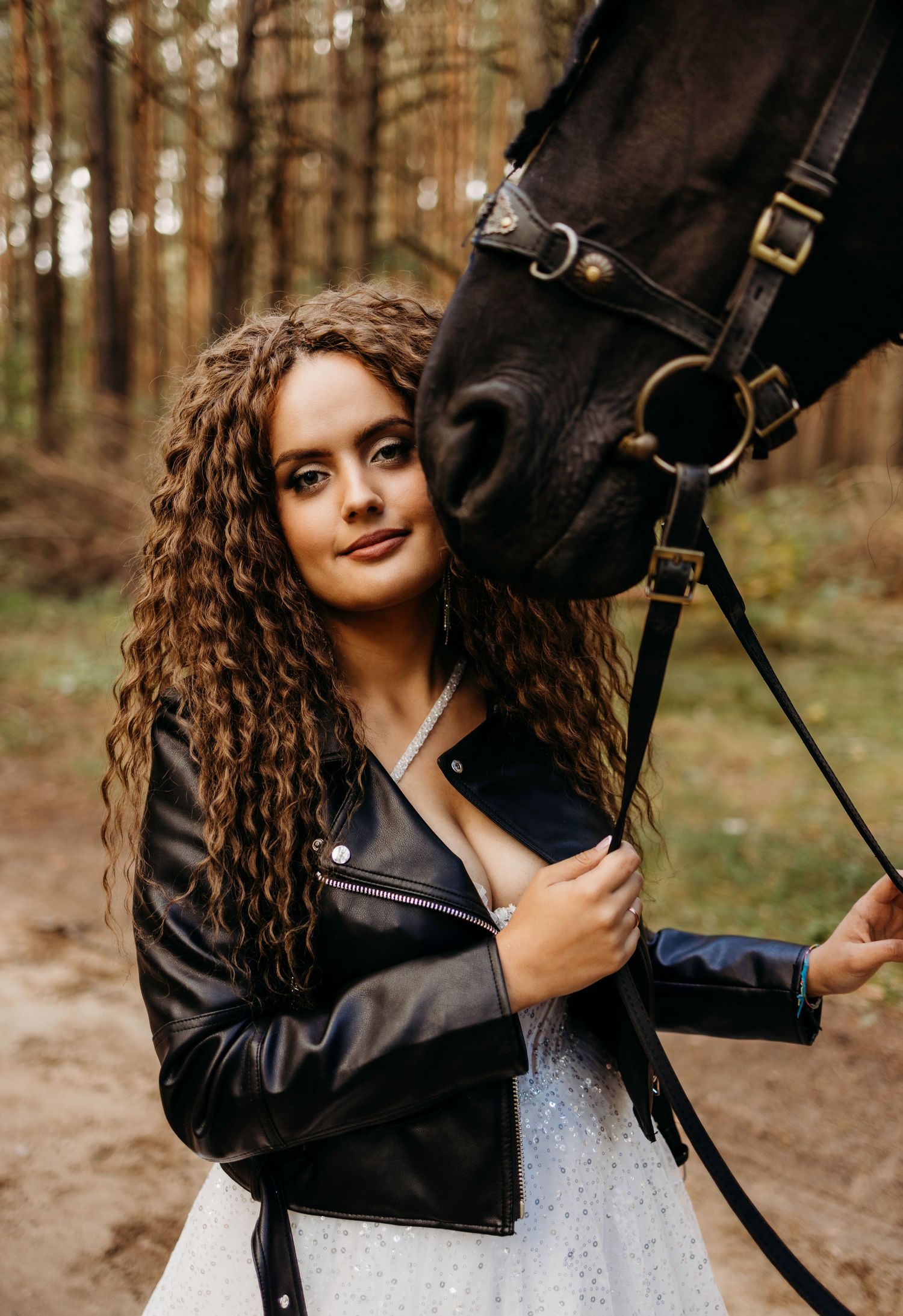 A person in a black leather jacket poses with a dark horse in a wooded forest setting.