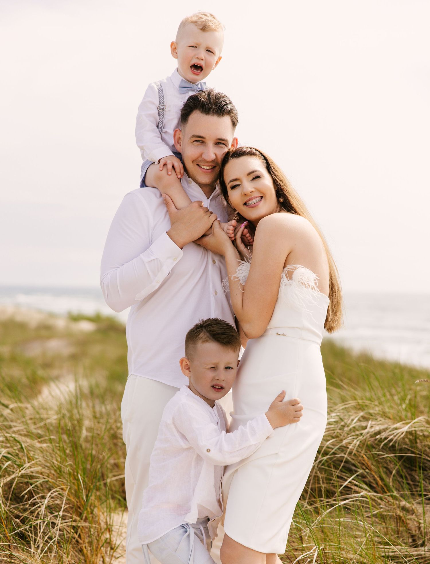 Family wearing white outfits embracing and laughing together in beach grass during sunset photoshoot.