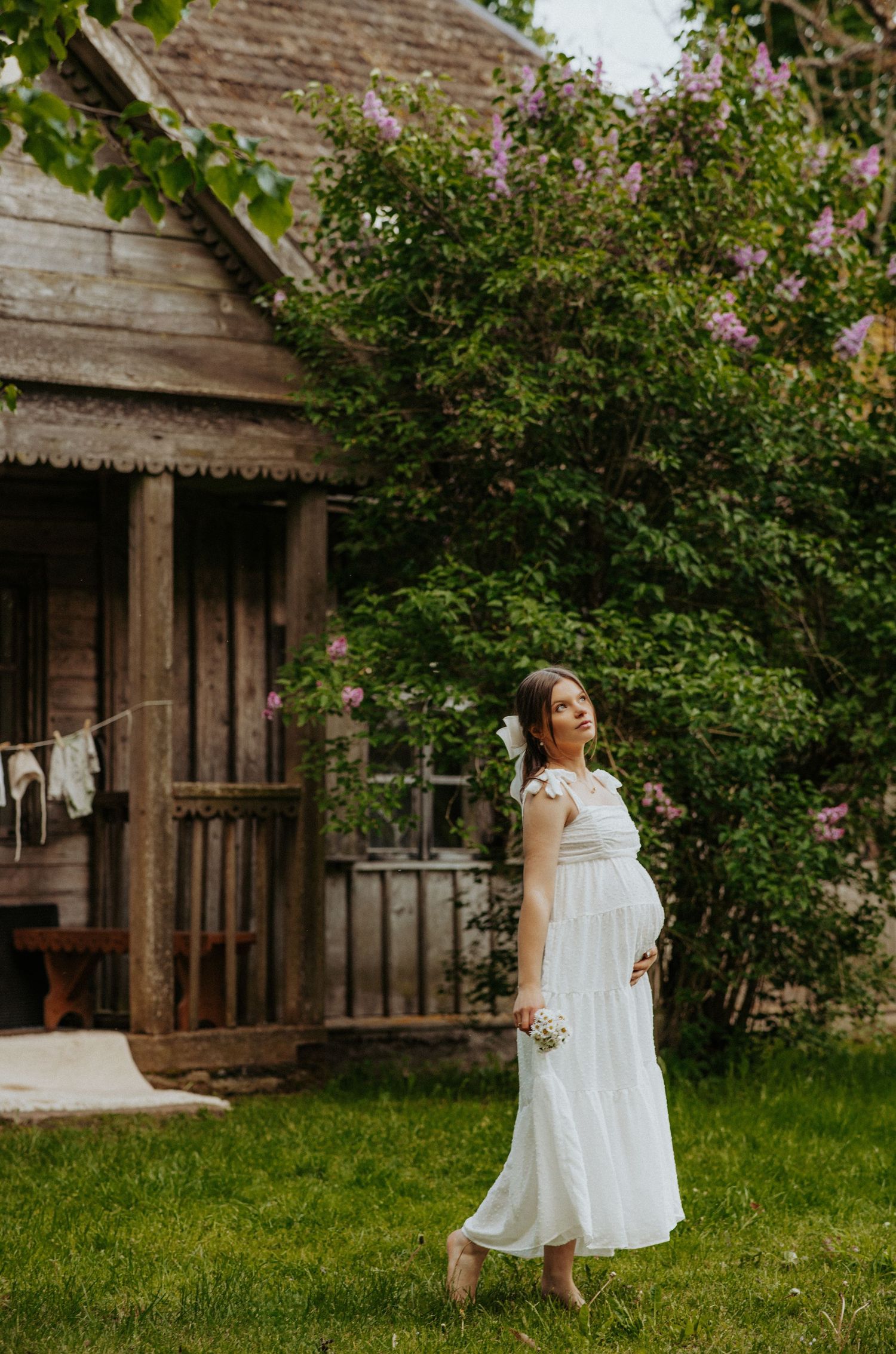 White dress maternity photos by rustic cabin surrounded by blooming spring foliage.