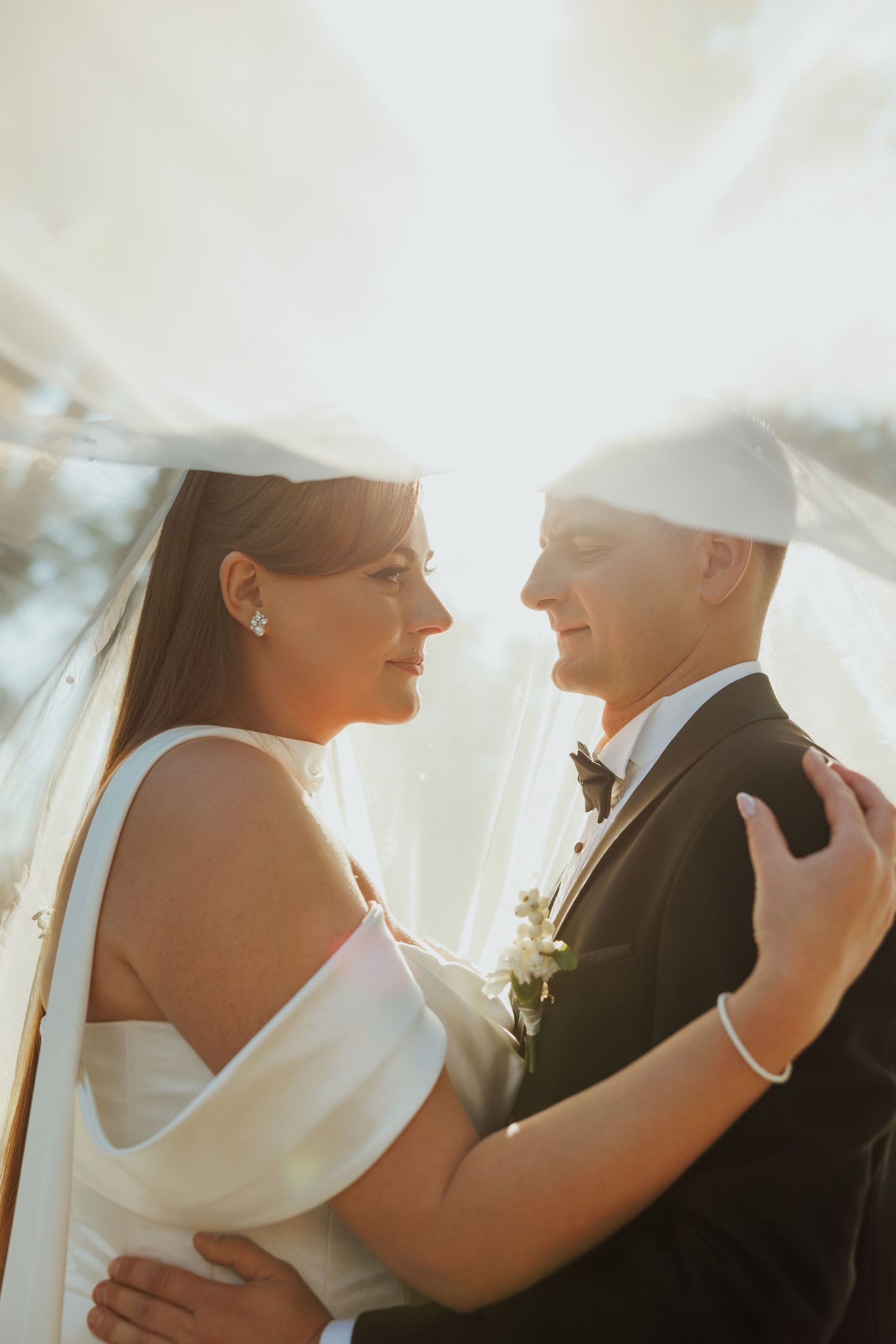 Intimate wedding couple portrait with soft sunlight and flowing white veil.
