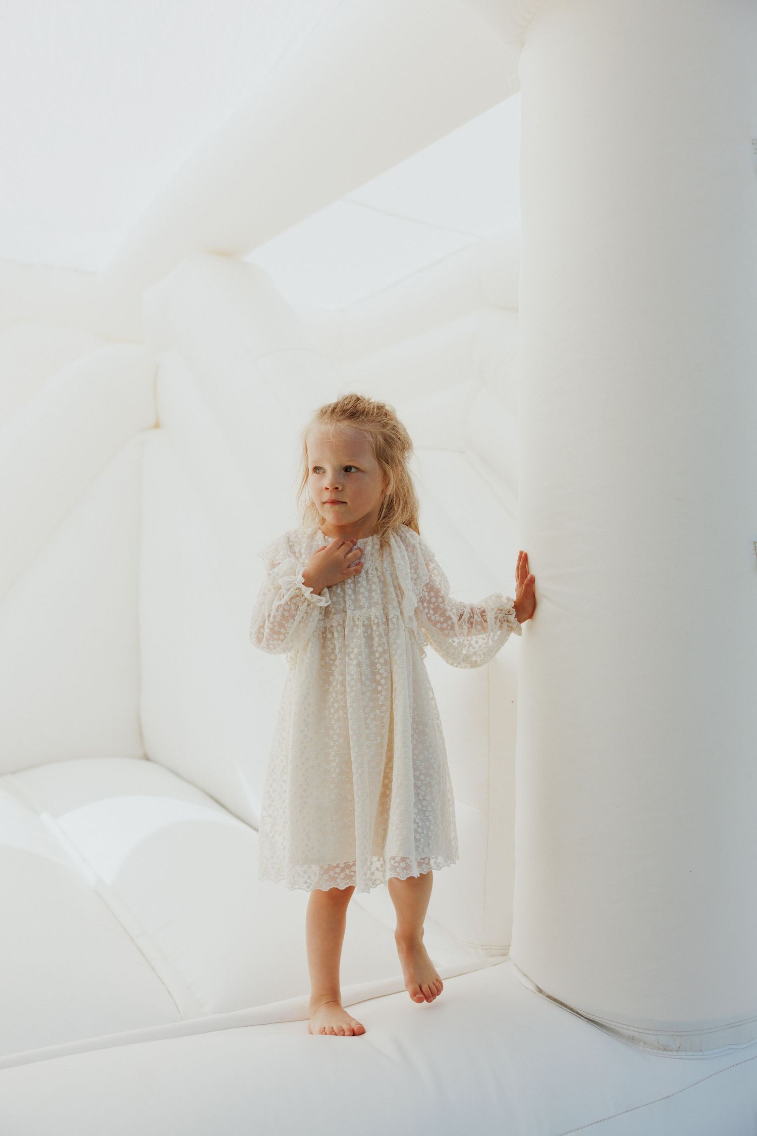 Small child wearing a delicate white dress standing on white surface.