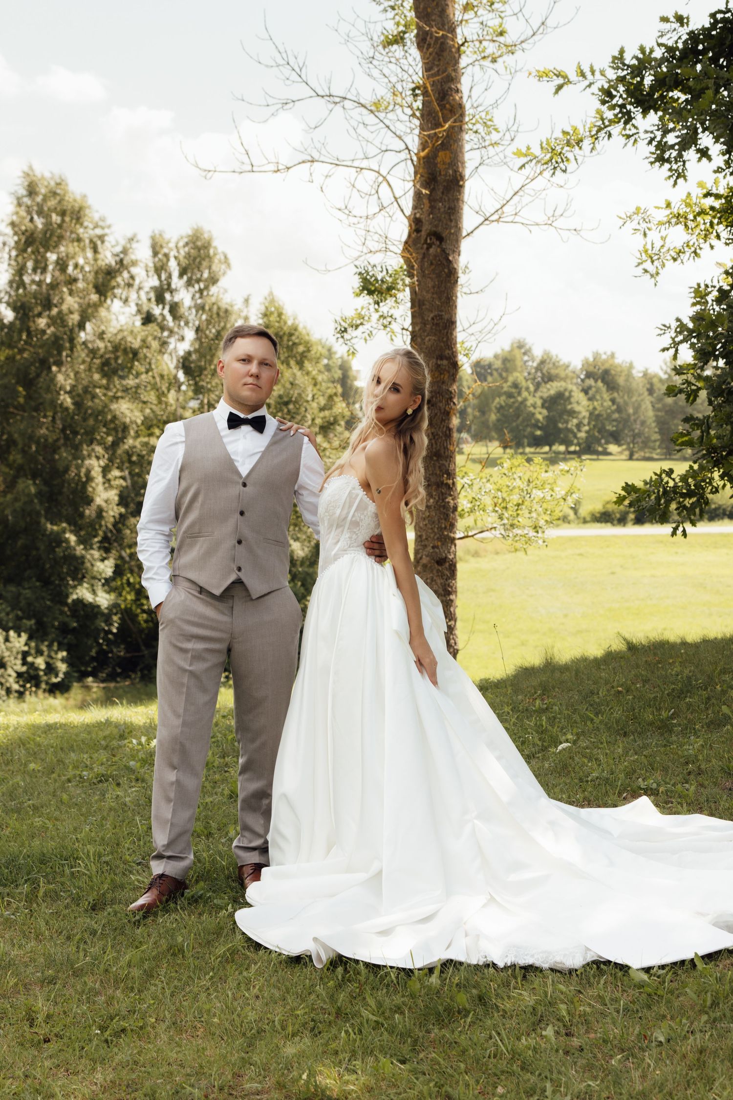 Bride in white gown and groom in gray vest with bow tie pose together outdoors near a tree on their wedding day.