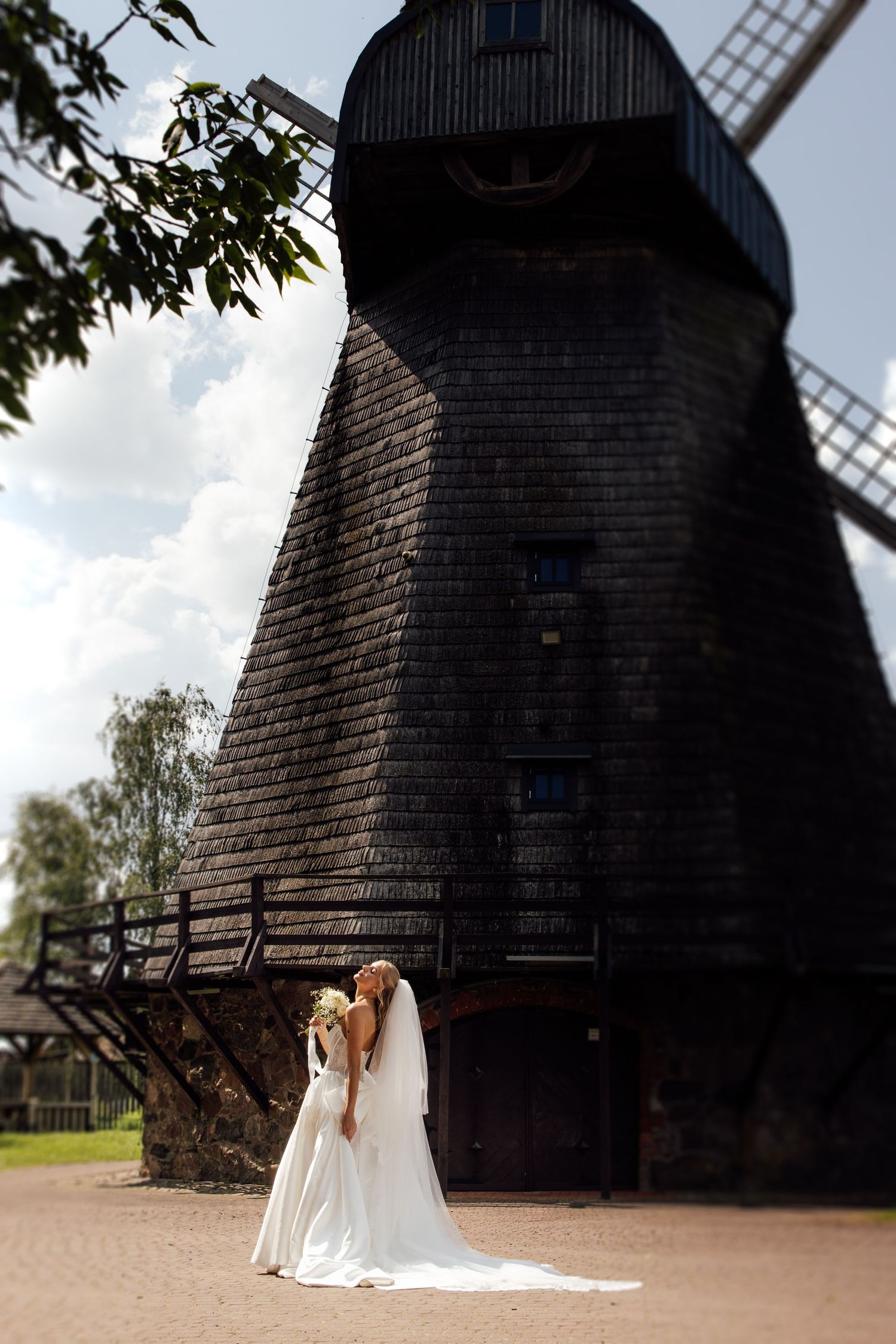 Bride in white gown and veil stands before a historic black wooden windmill under a cloudy sky.