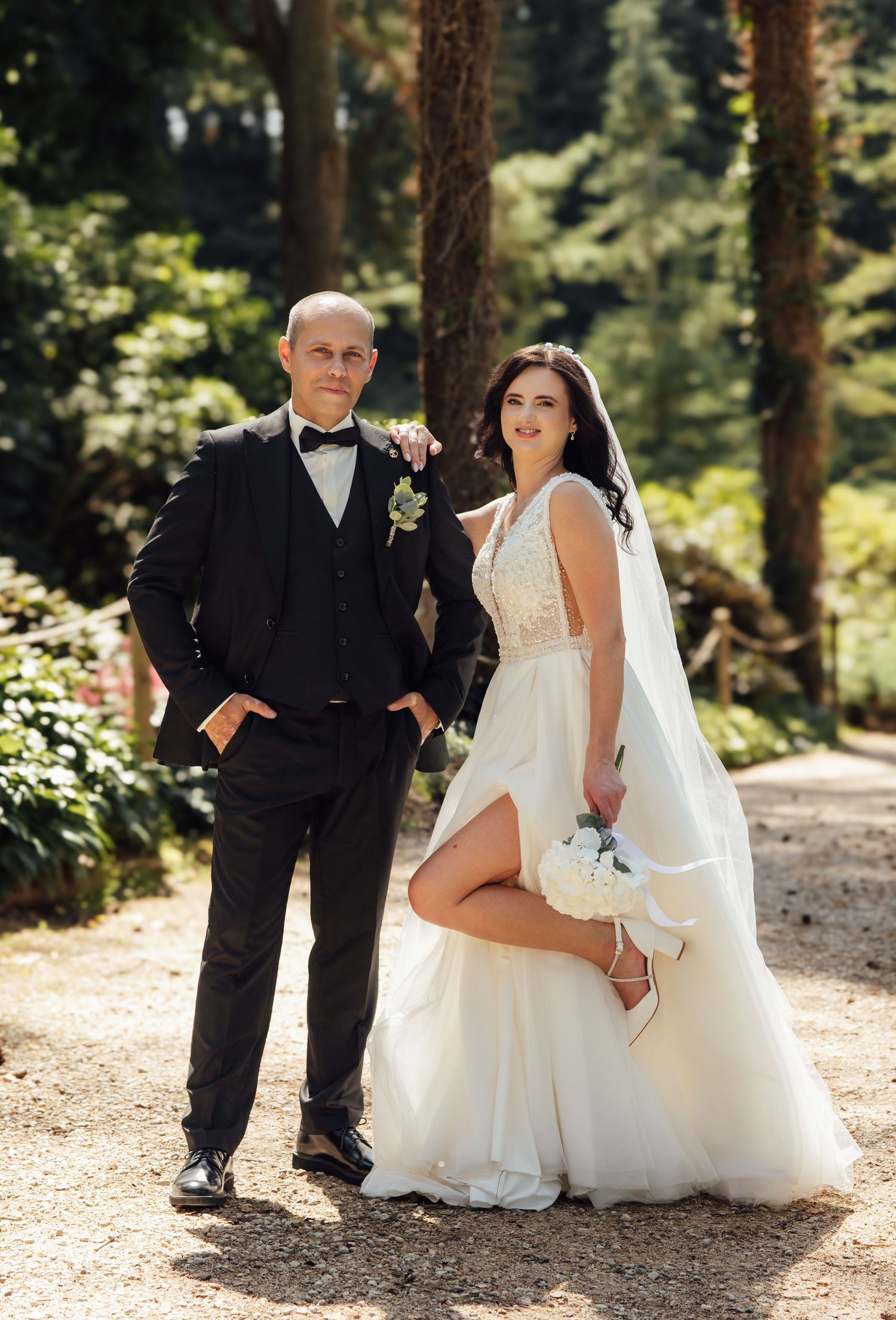 Elegant wedding couple posing in sunlit garden, groom in black tuxedo and bride in white dress with slit showing heels.
