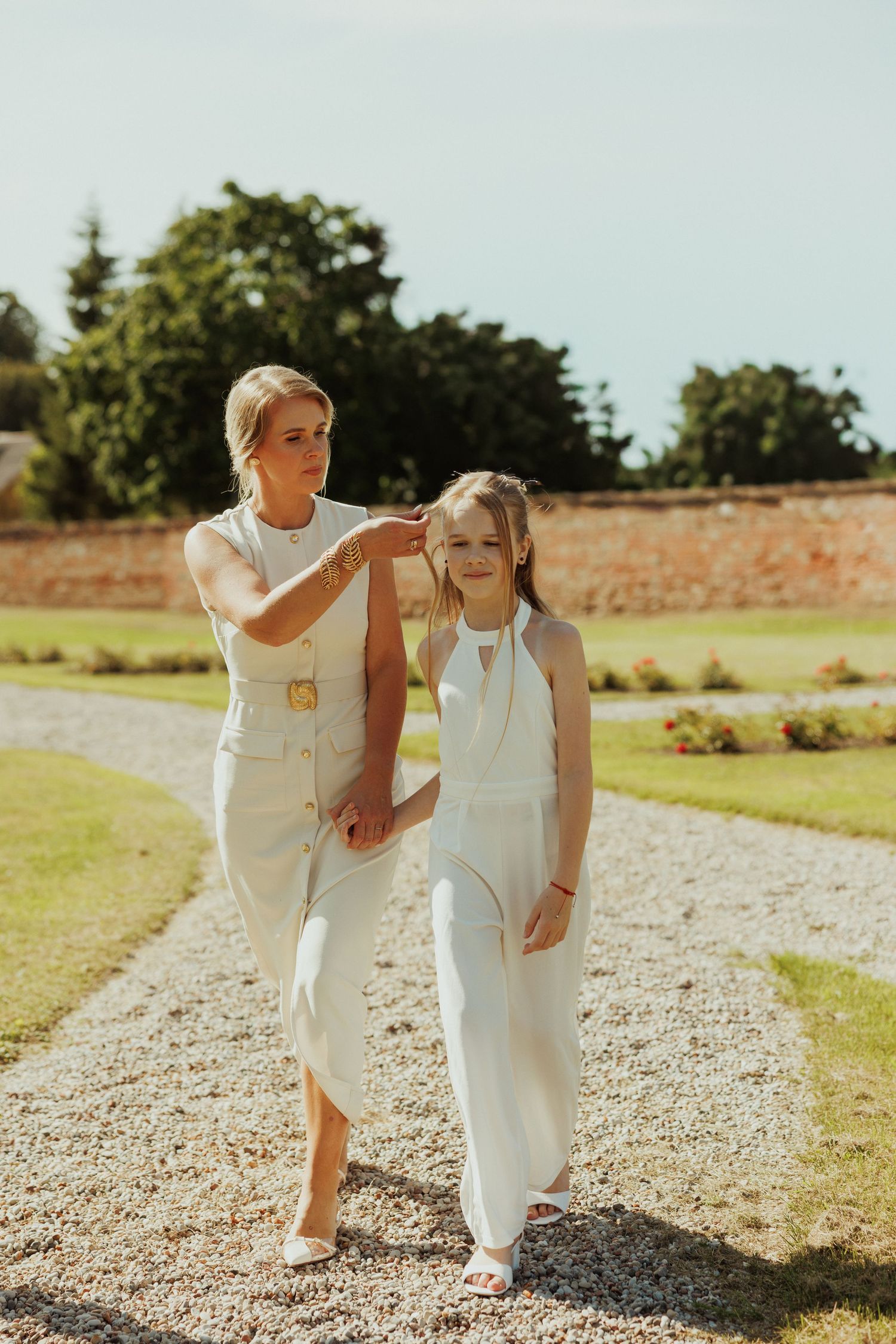 Two people in coordinating white outfits stand together on a gravel path in a sunny garden.