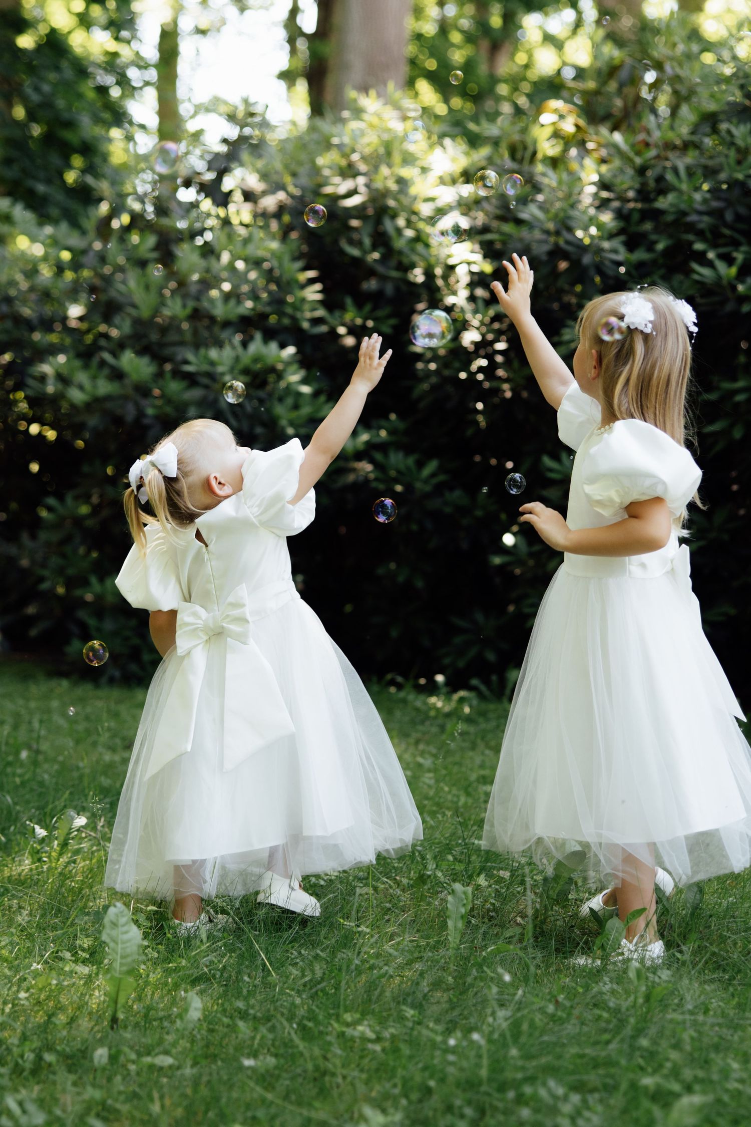 Two little girls in white tulle dresses reach up to catch soap bubbles in a lush green garden.