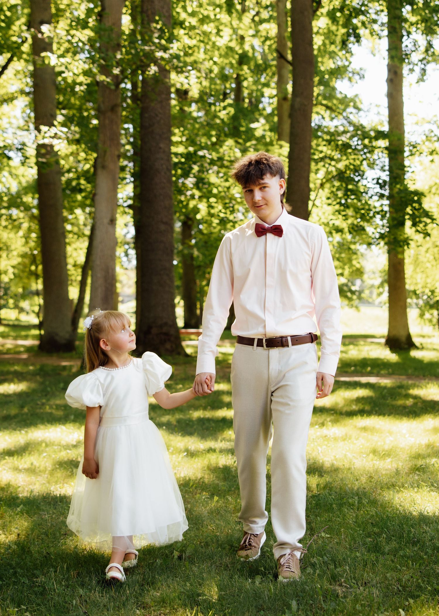 A teenage boy in a pink shirt and bow tie holds hands with a young girl in a white dress in a sunny park.