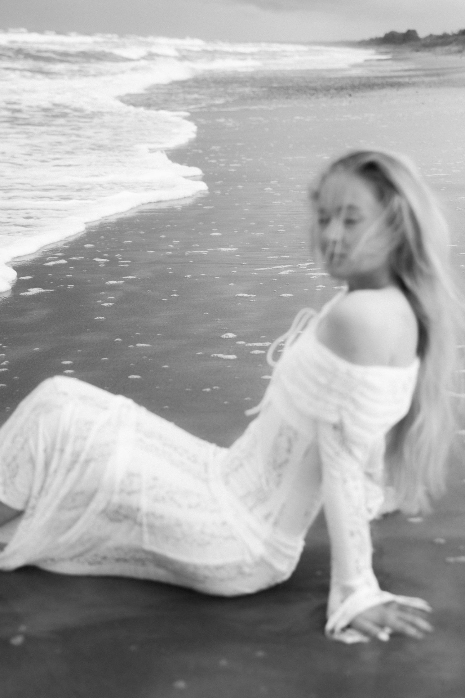 Black and white photo of a woman in a white lace dress sitting on a beach shore with waves nearby.