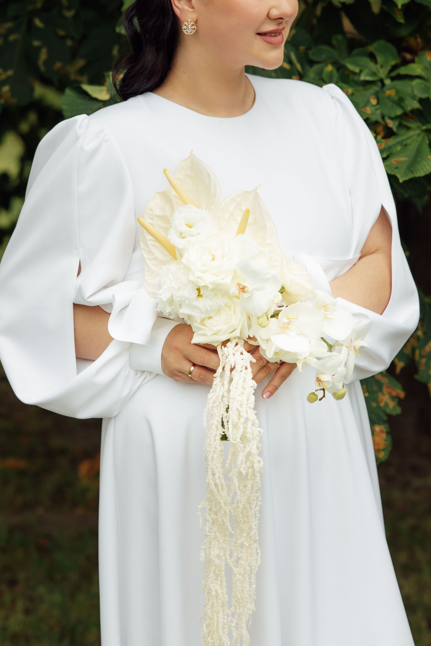 Bride in white long-sleeve gown holding elegant ivory floral bouquet with trailing pearl accents, standing outdoors.