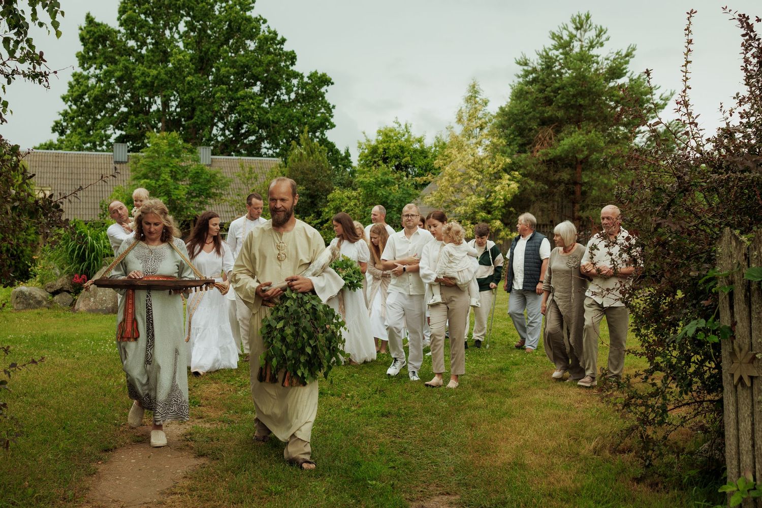 Group of people in white attire walking in a garden procession, carrying greenery in an outdoor ceremony.