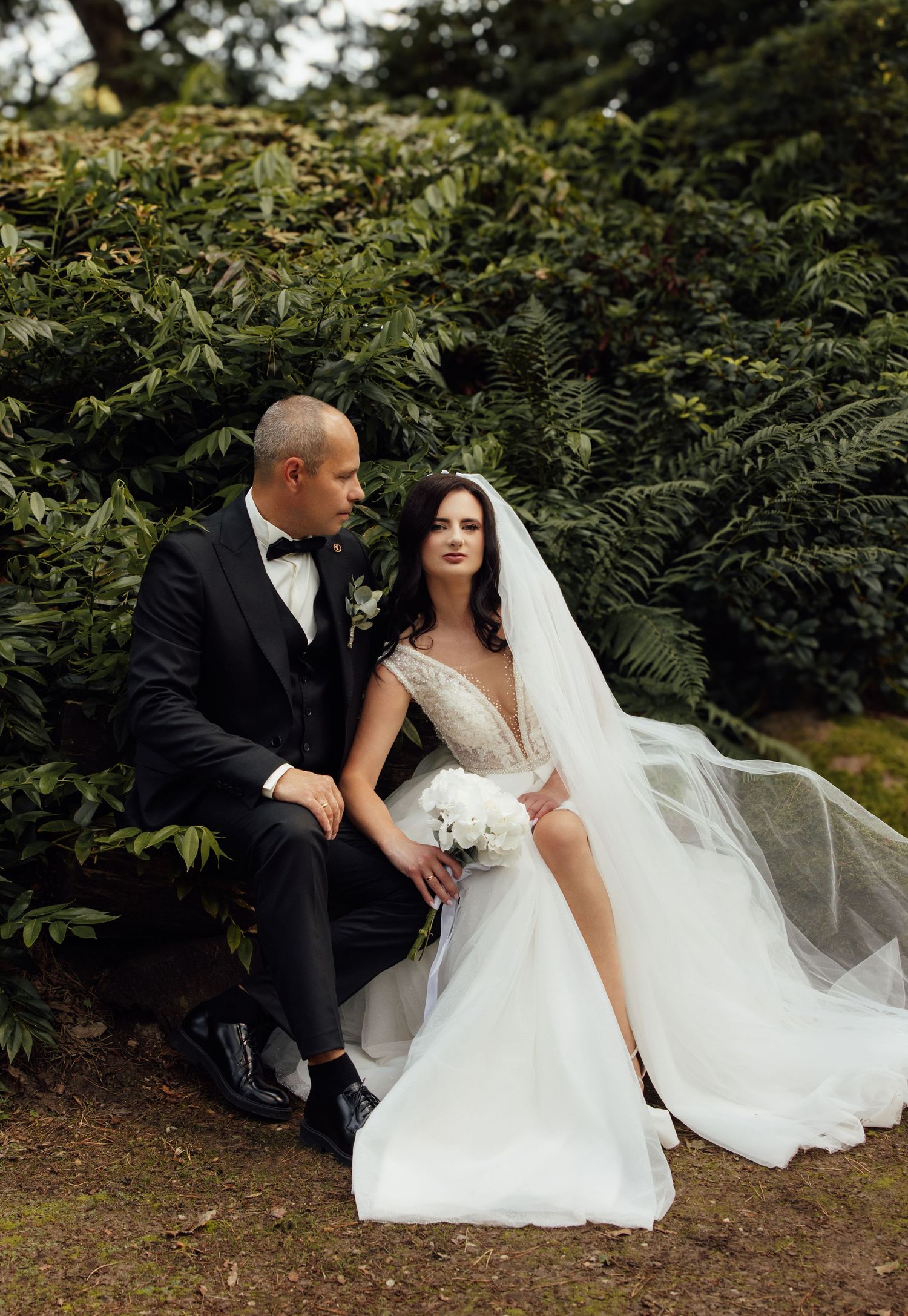 Bride in white gown with veil and groom in black tuxedo sitting together in lush green garden.