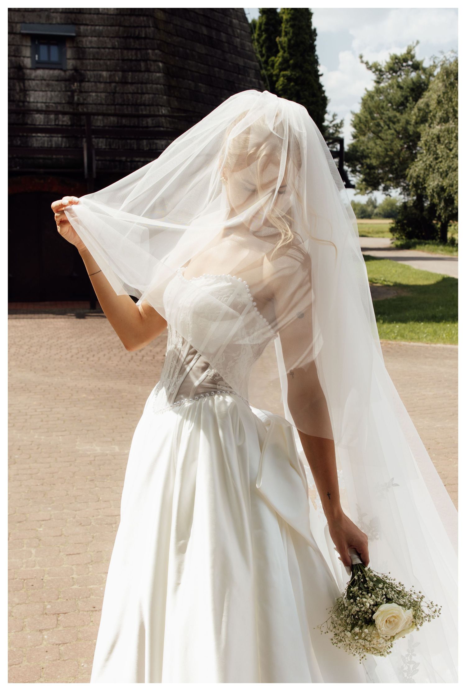 Bride in white gown holding veil over face while clutching bouquet outdoors near rustic barn.
