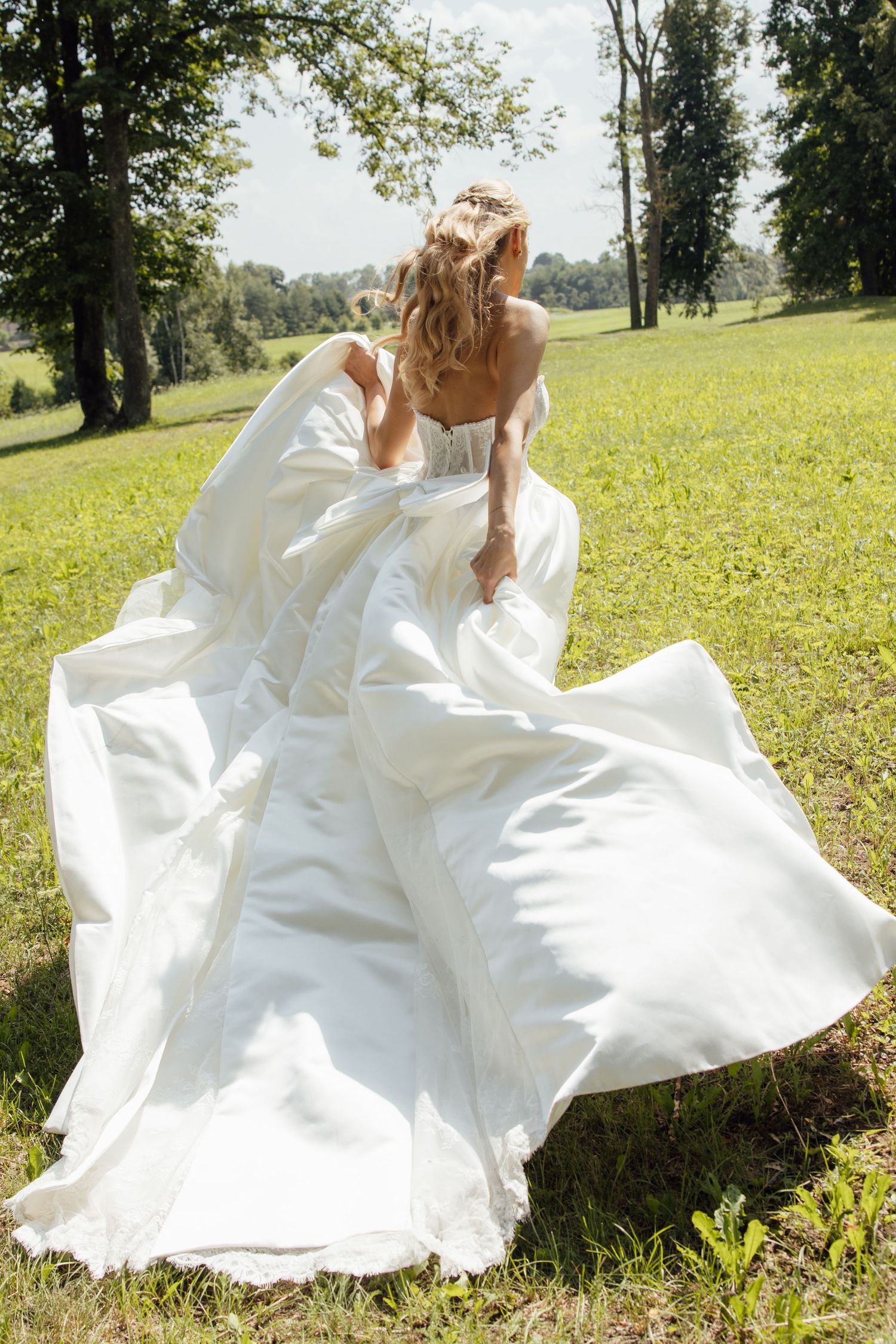 Bride in flowing white wedding dress running through green meadow with trees, dress billowing in the wind.
