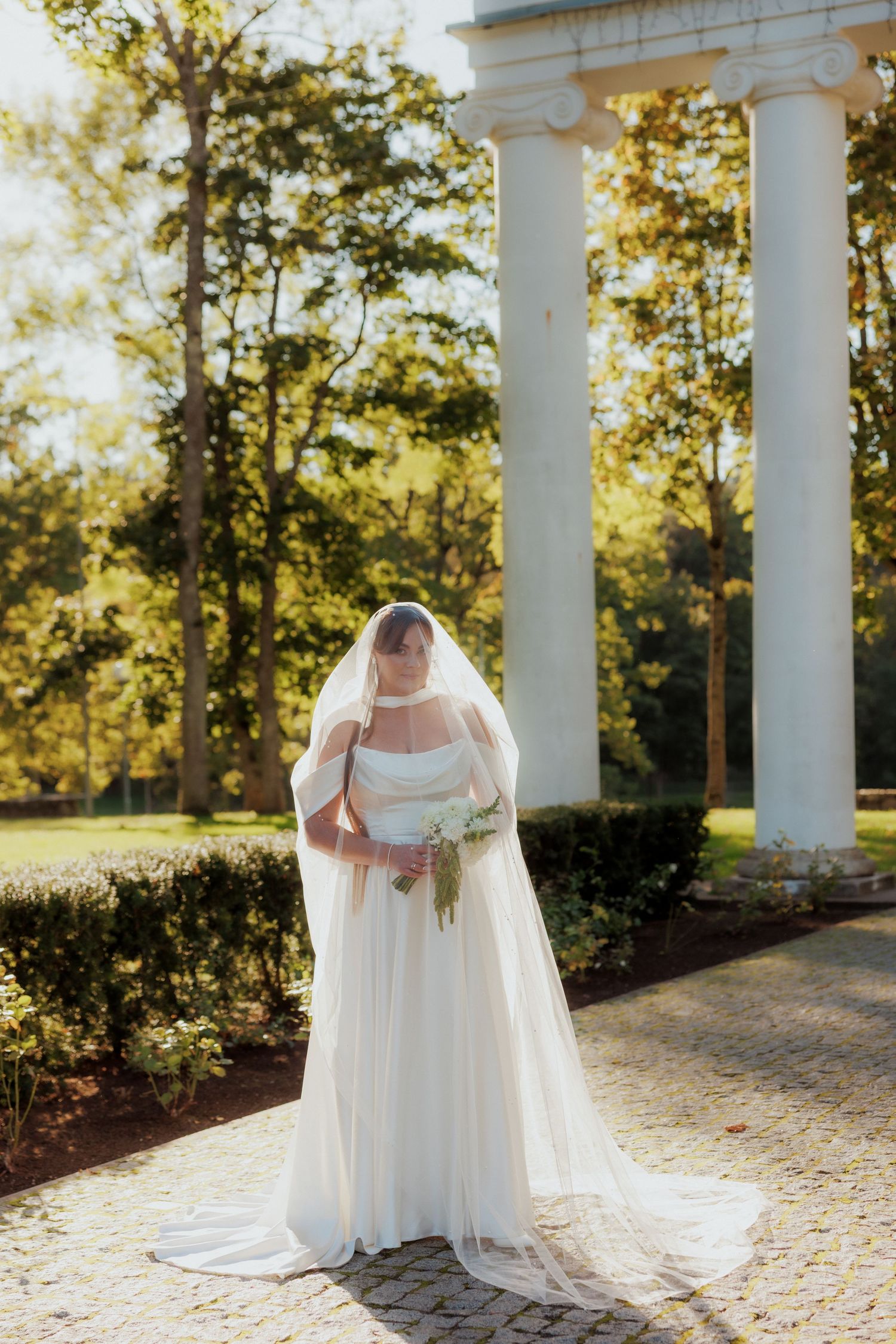 Bride in flowing white dress poses among classical white columns in golden sunlight.