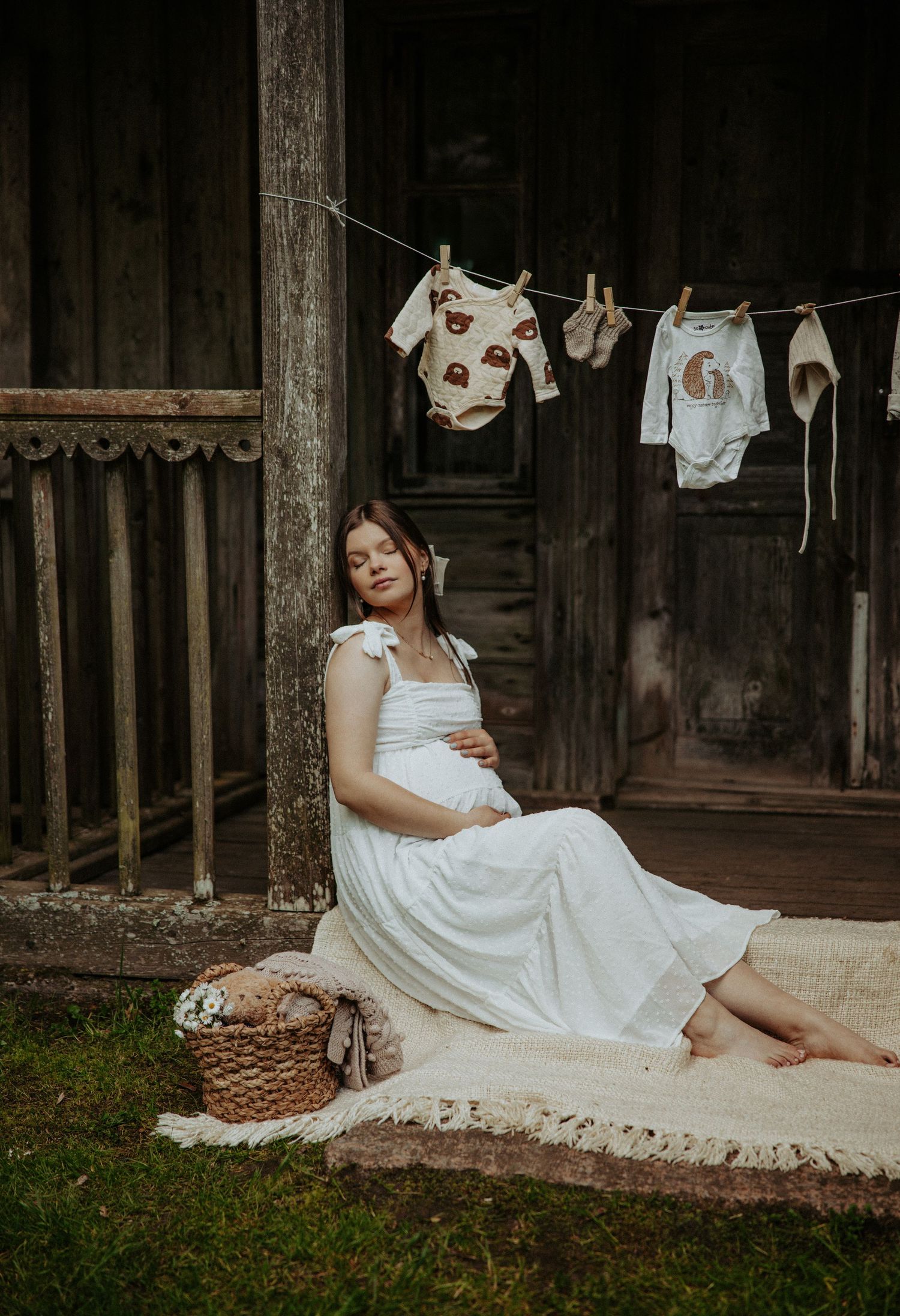 Expectant figure in white dress sits by rustic barn wall with baby clothes on display.