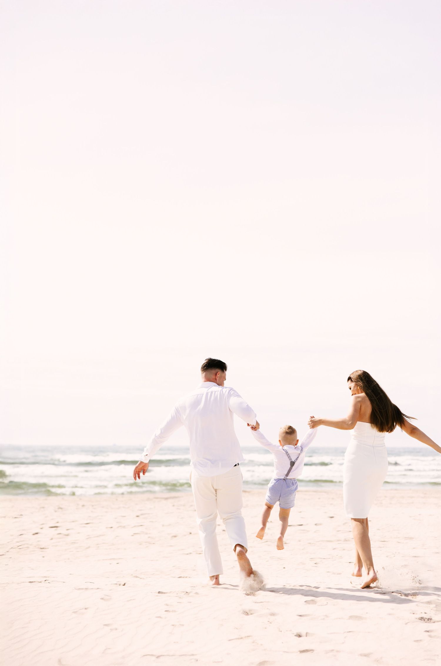 Family in white outfits running and playing together on sunny beach.