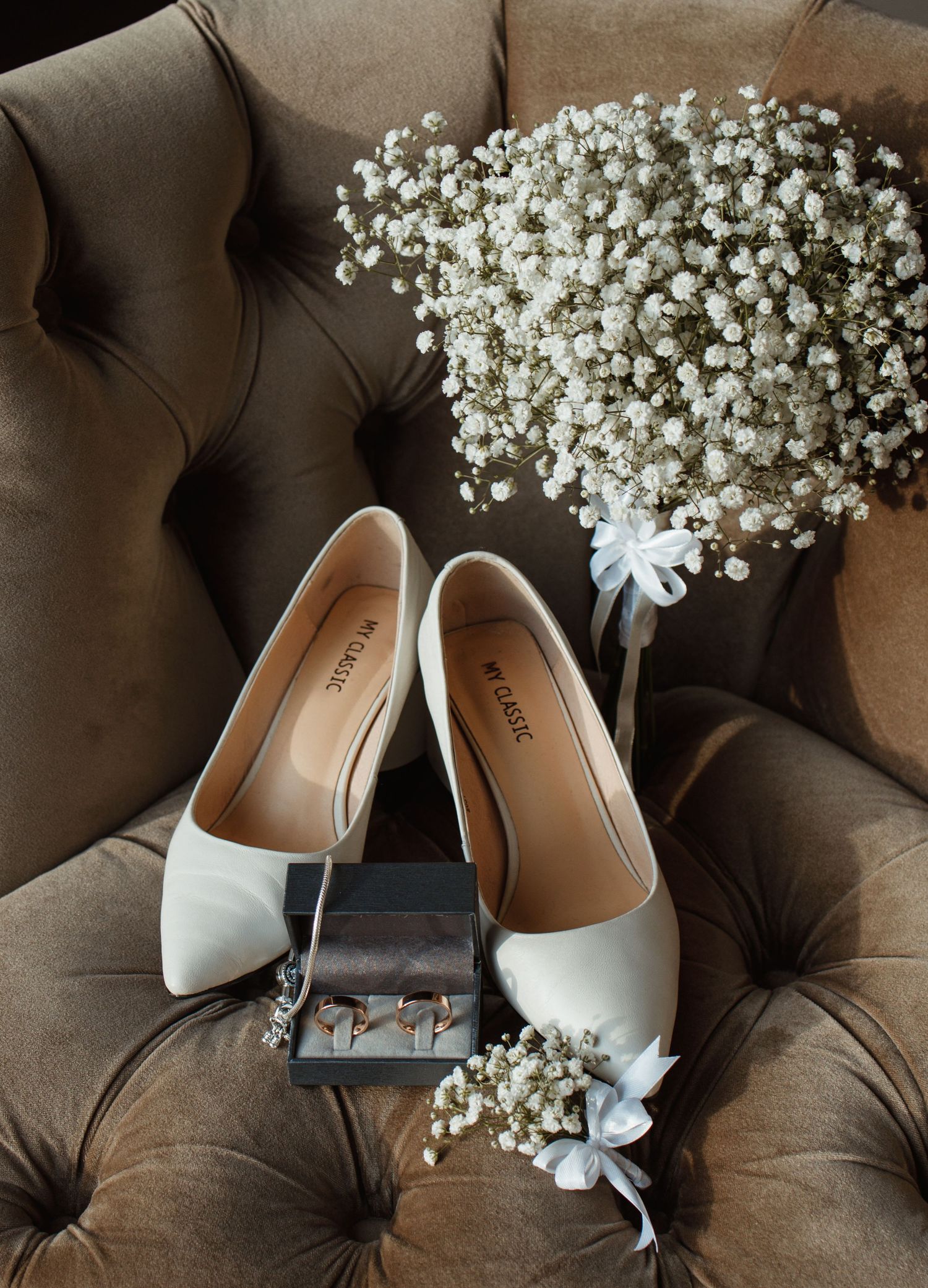 White bridal shoes and baby's breath flowers arranged on brown tufted chair.