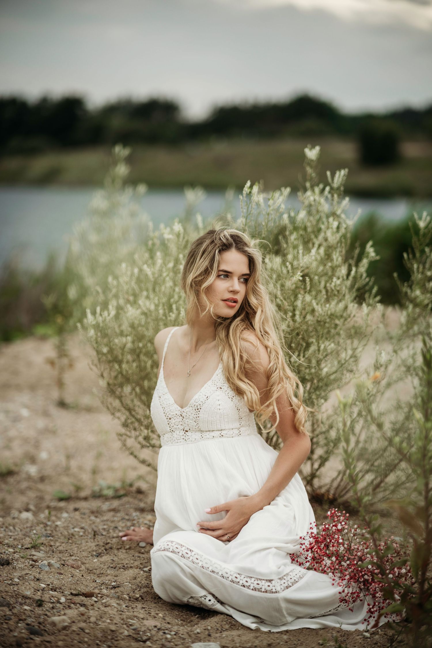 Woman in white lace dress poses elegantly among coastal vegetation.