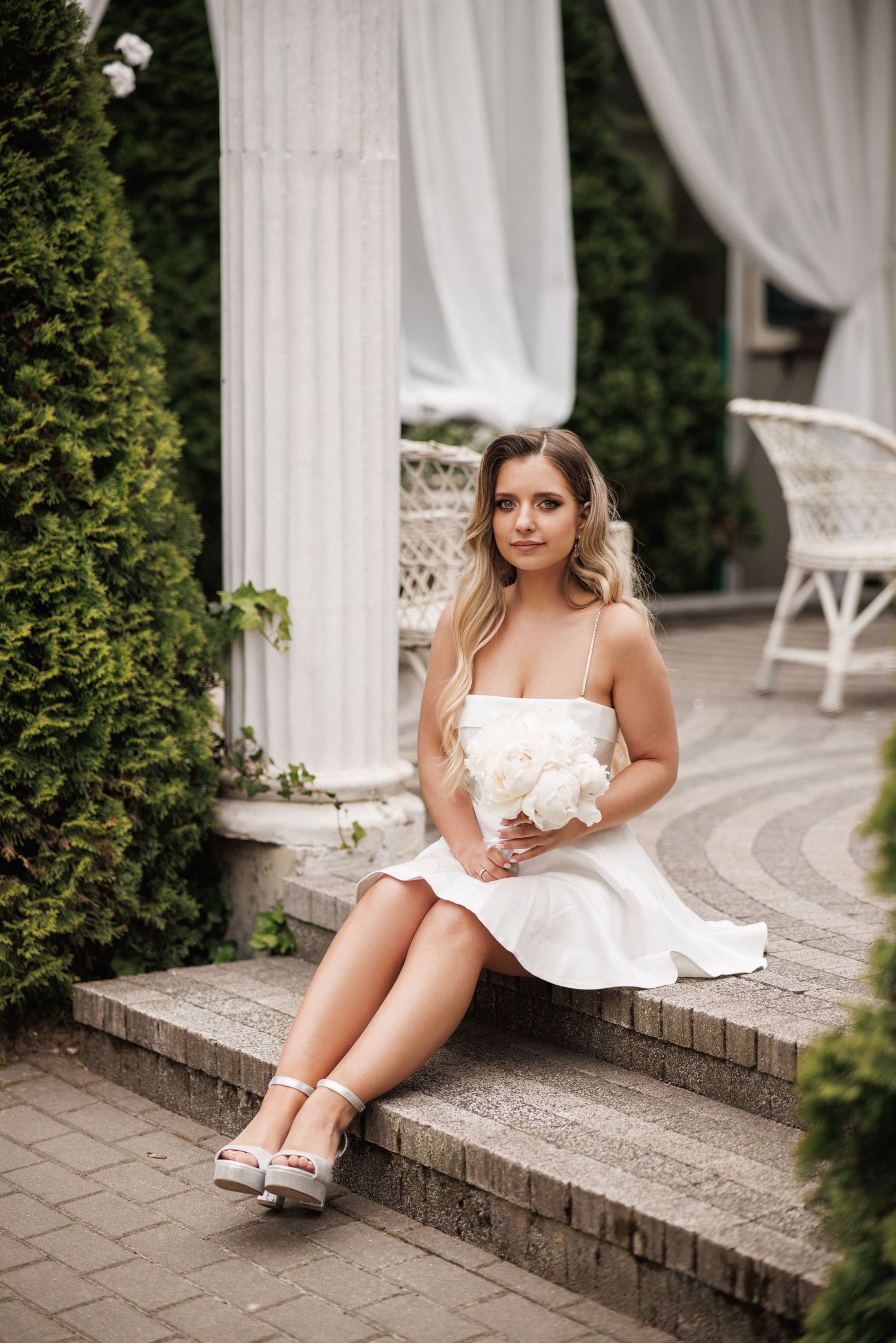 A woman in a white dress sits elegantly on stone steps near white curtains and greenery in an outdoor setting.
