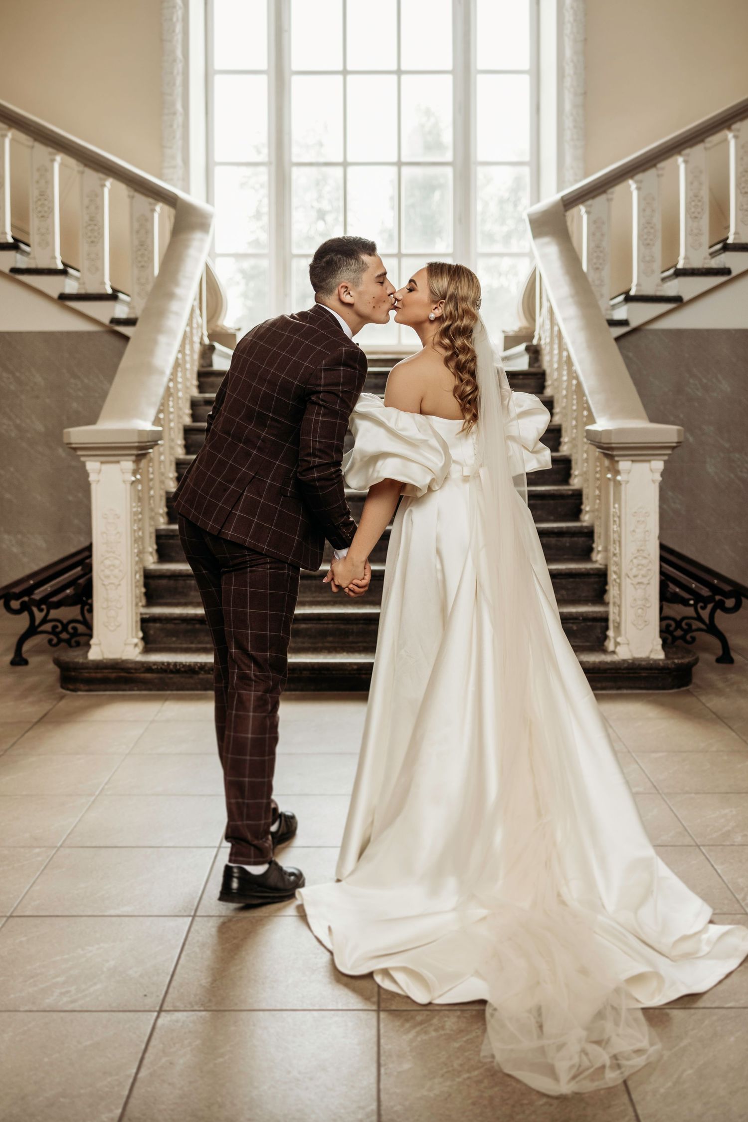 A romantic wedding couple sharing a kiss on an elegant white staircase, with the groom in plaid and bride in flowing gown.
