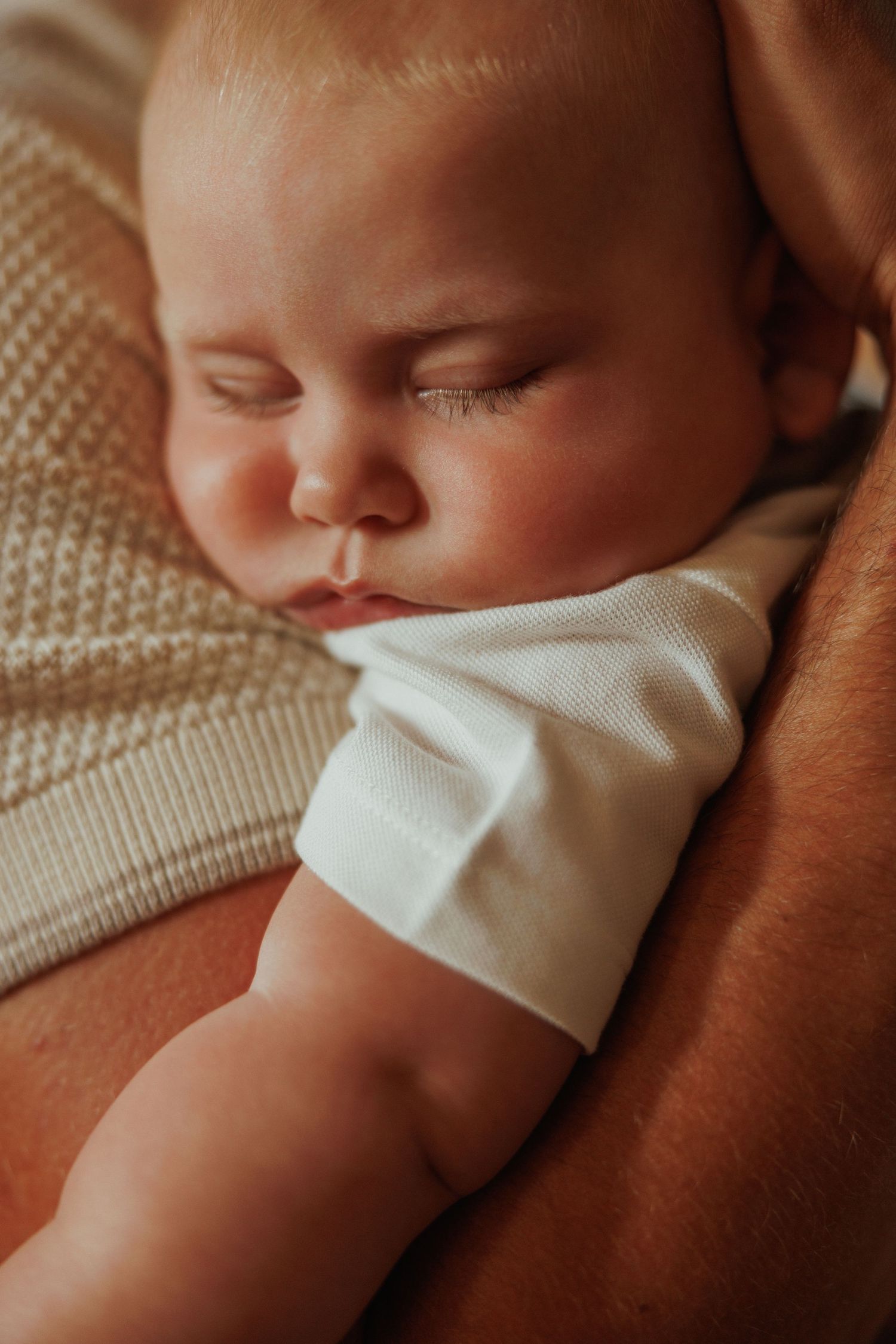 A sleeping baby in white clothing rests peacefully on a brown blanket in a close-up portrait.