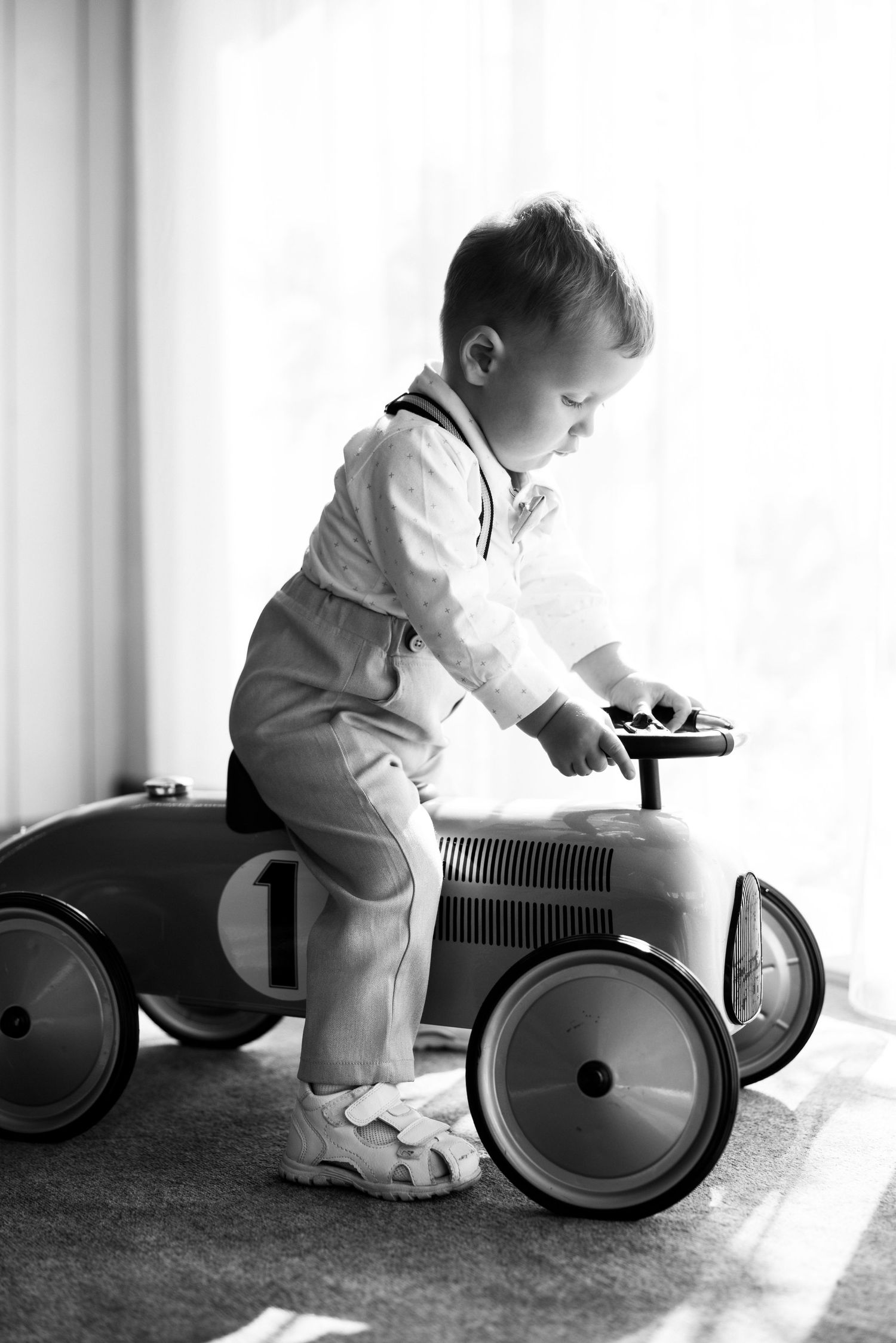Black and white photo series showing a small child playing with a vintage ride-on toy car in natural window light.