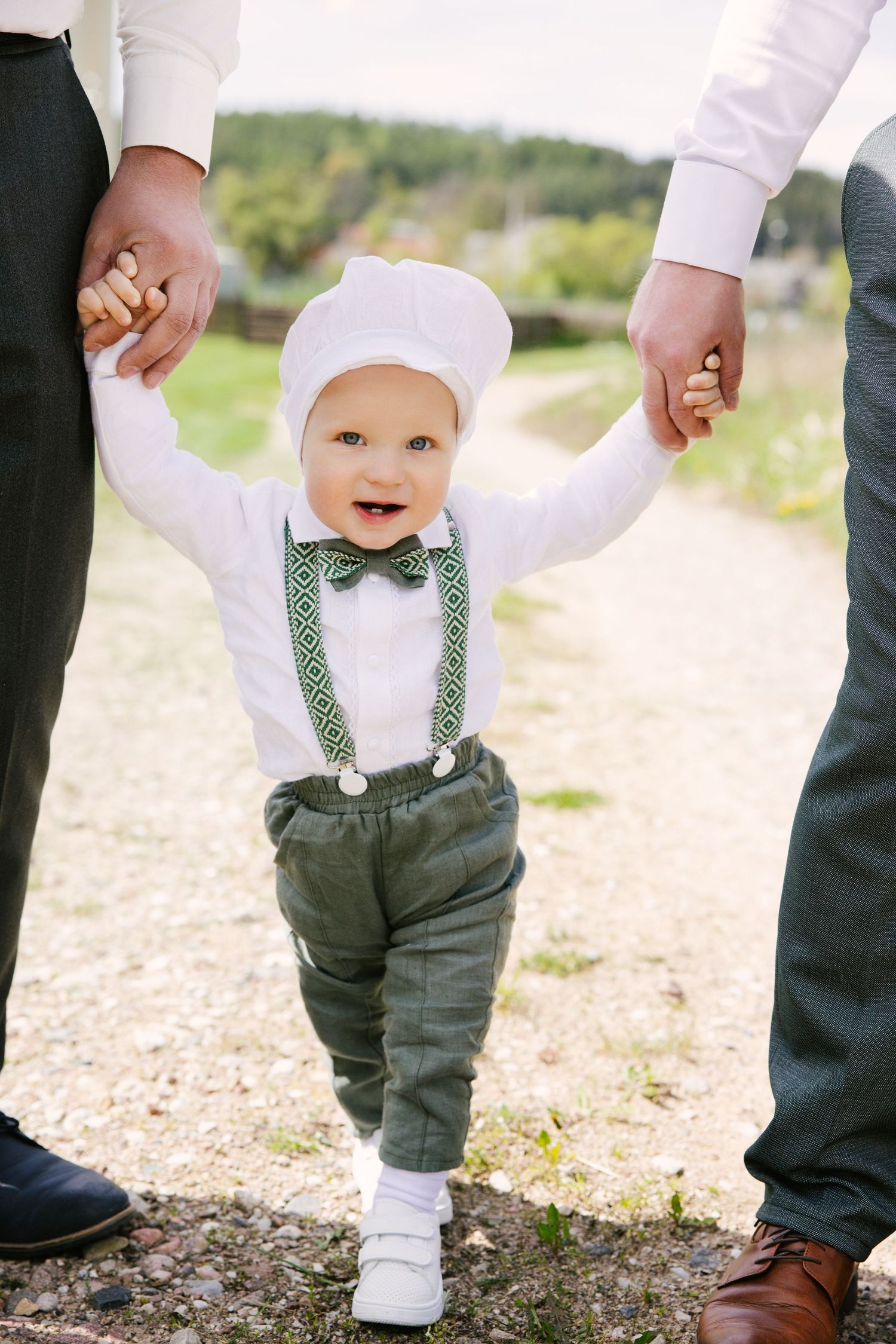 Close-up of tiny feet in white shoes and grey suspender pants walking with adult guidance.