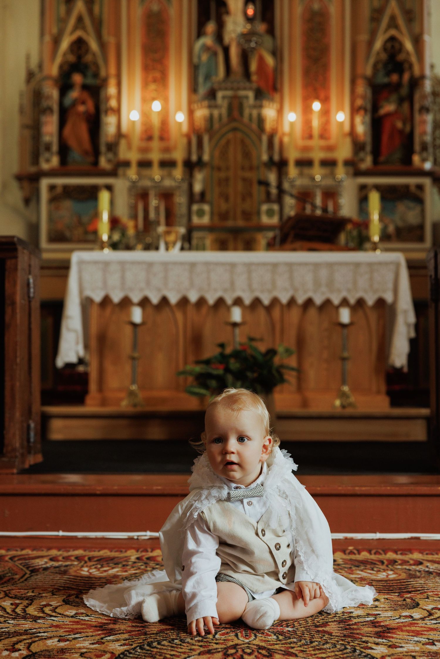 Baby in white christening gown sits on ornate carpet in front of church altar with glowing candles.