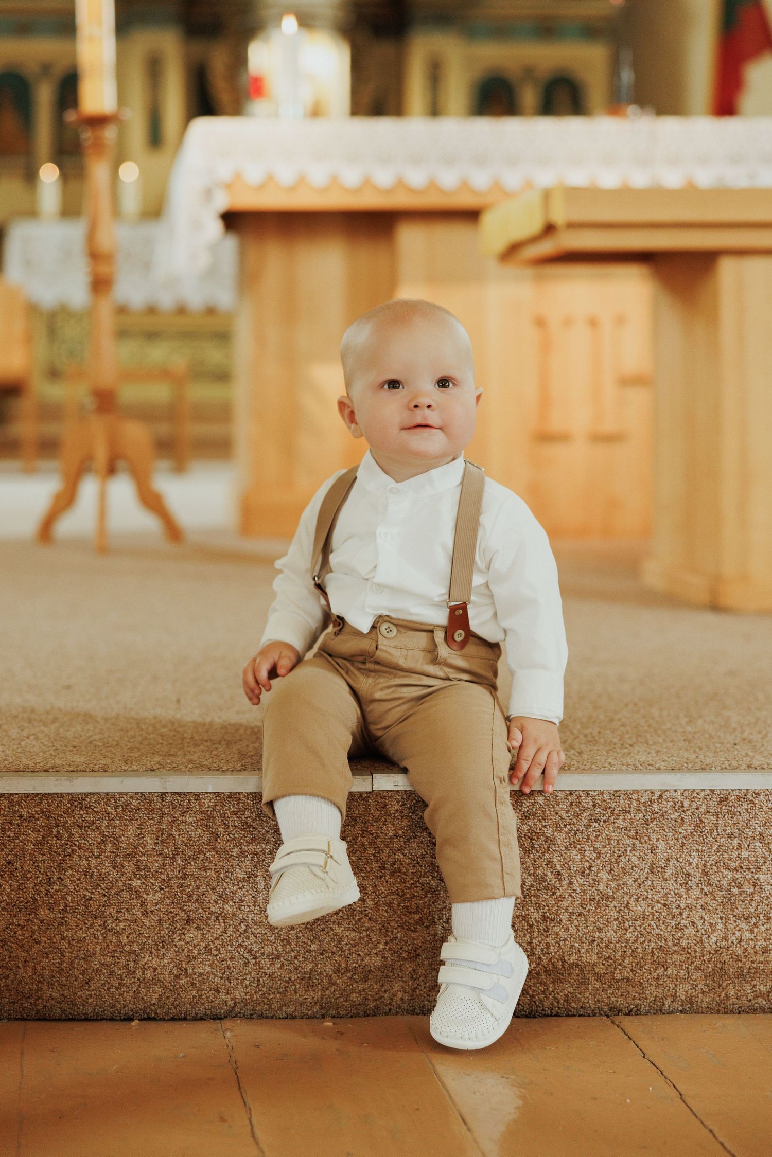 A toddler in white and brown clothing sits on carpeted steps in a warm-lit interior setting.