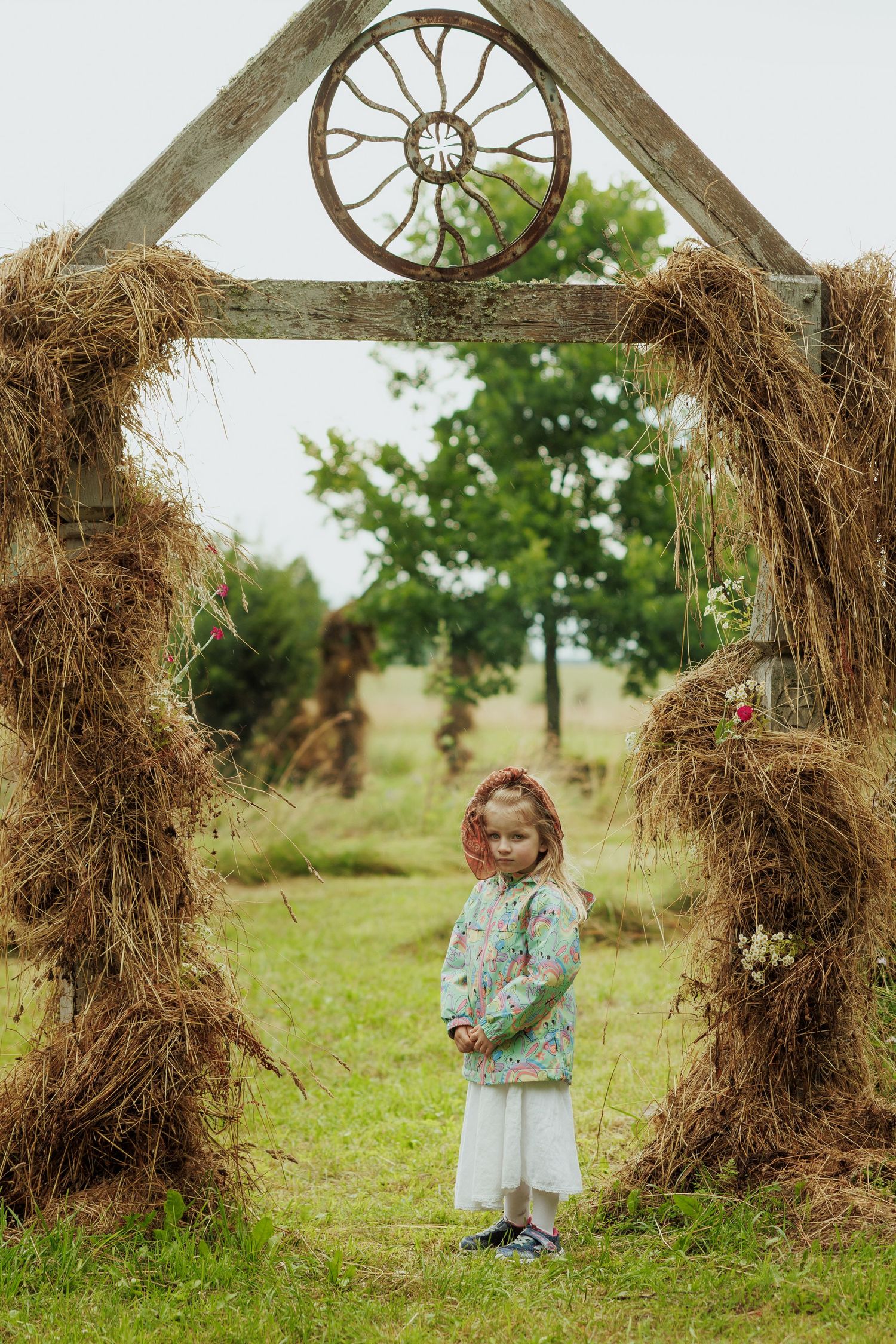 Young girl in floral jacket standing beneath rustic hay bale archway with wagon wheel decoration.