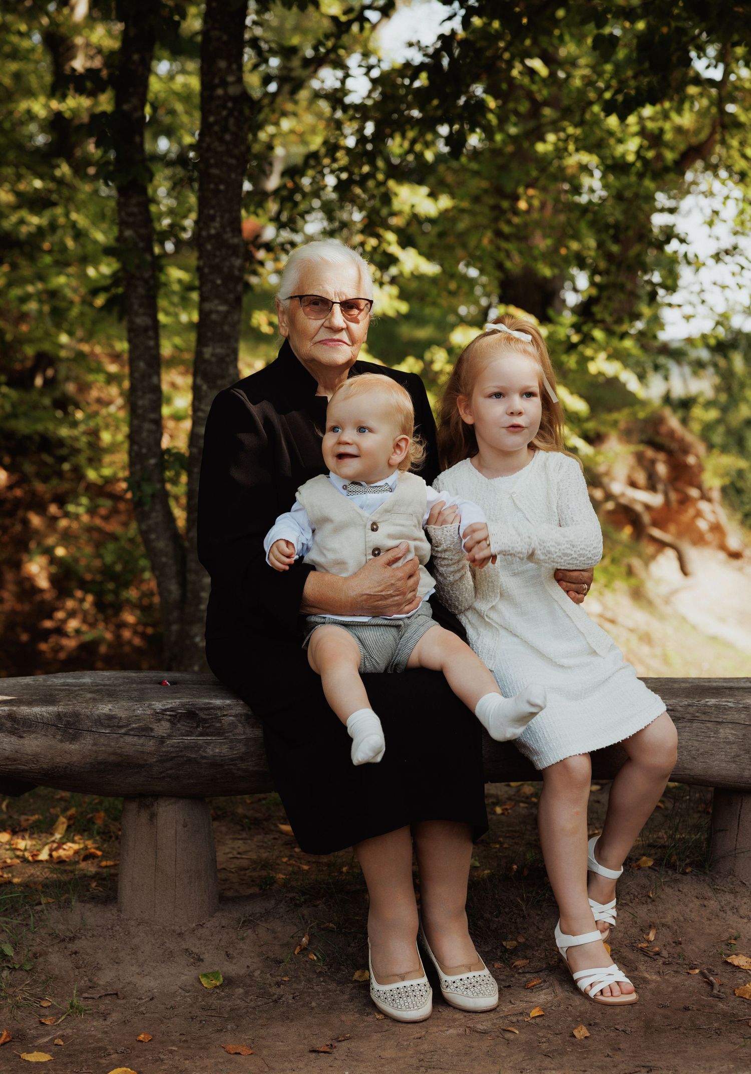 Two small children sit on a wooden bench in a sunlit outdoor setting wearing white outfits.