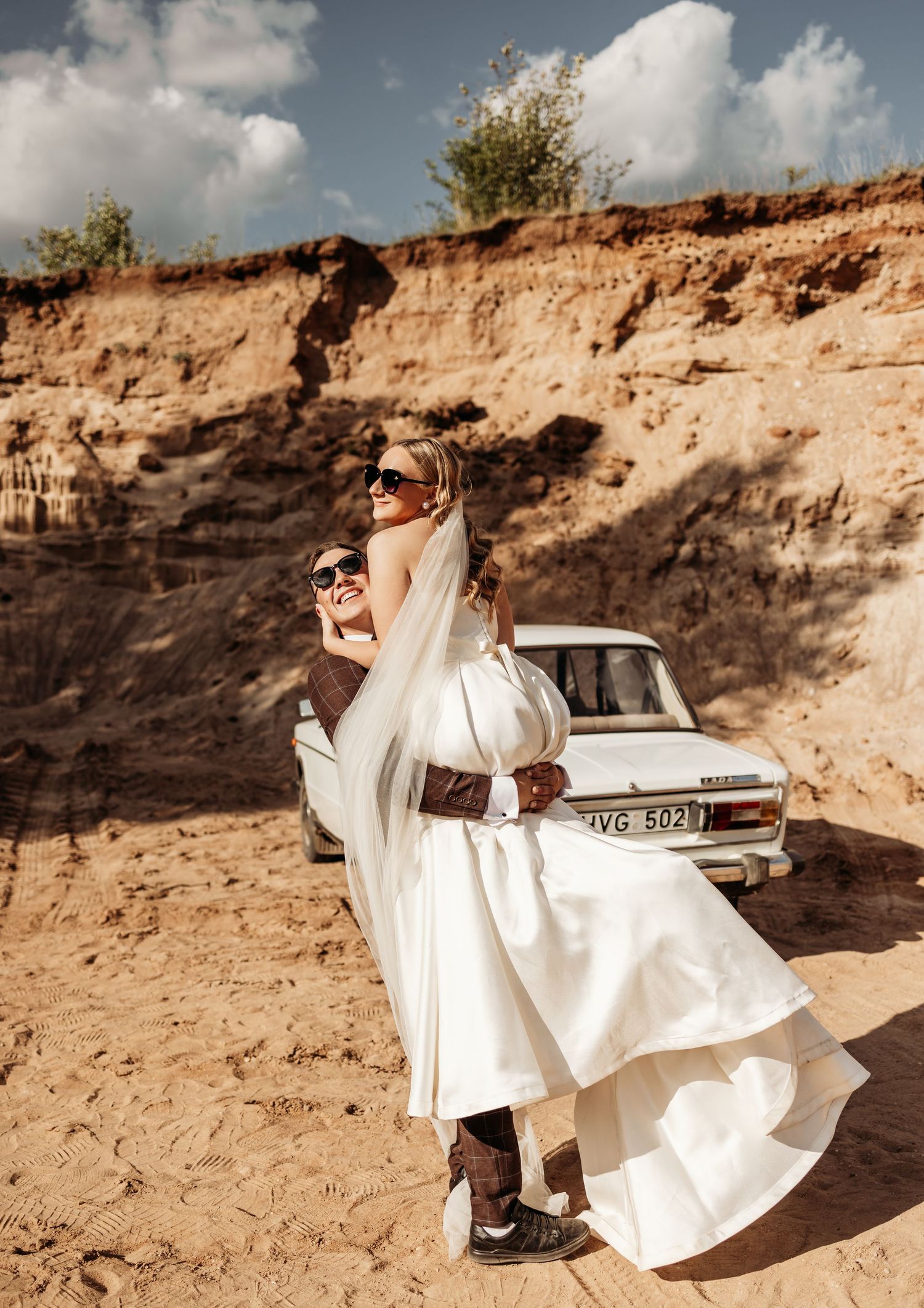 A person in a flowing white wedding dress poses dramatically against sandy cliffs with a vintage car in the background.