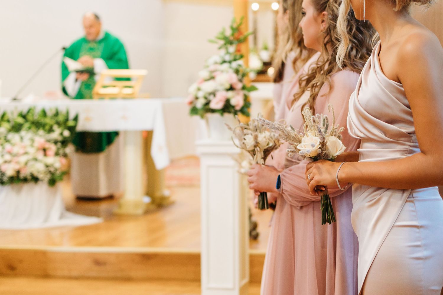Wedding party members in blush pink dresses hold elegant white and green floral bouquets during a formal ceremony.