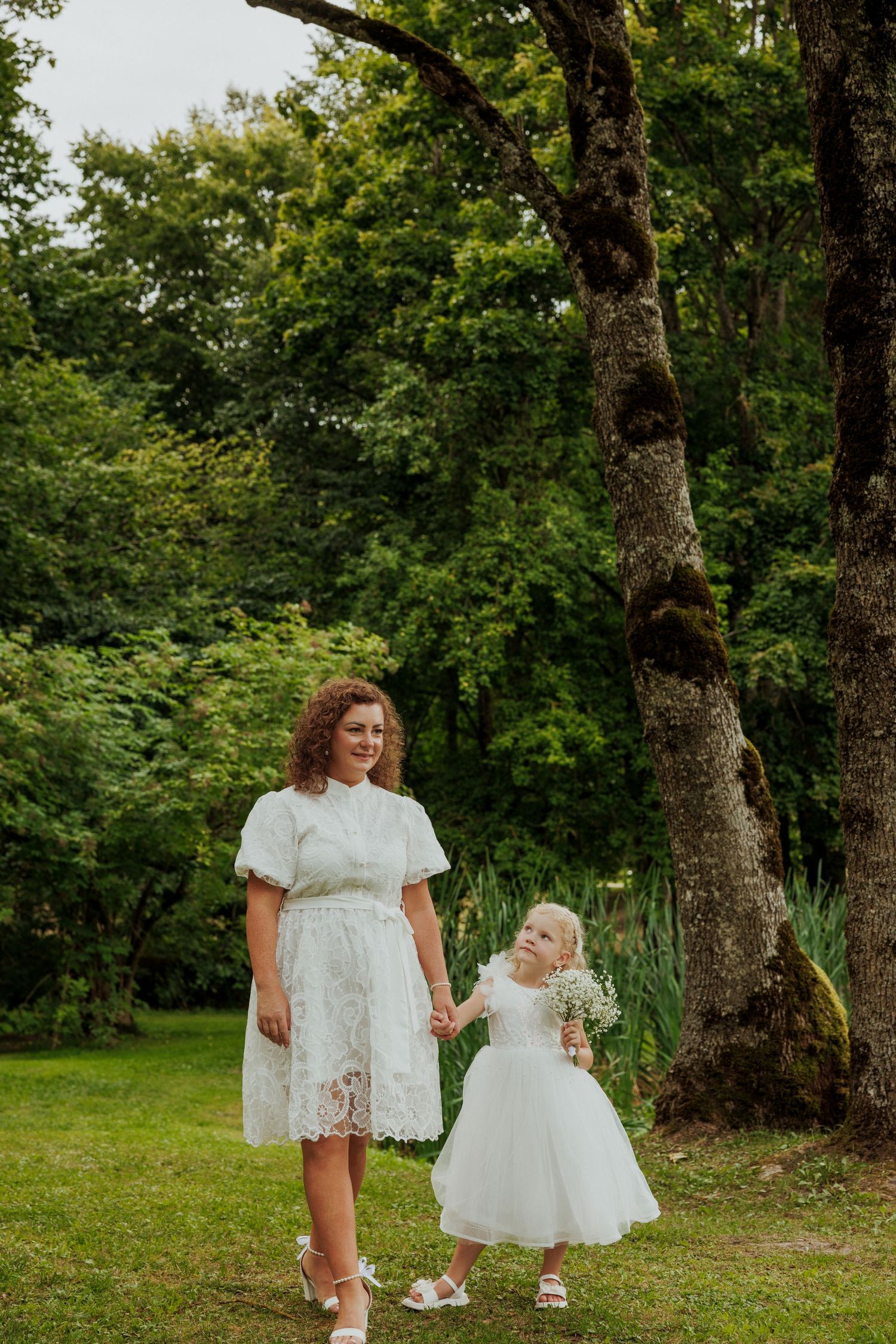 Two children in white lace dresses walk together on green grass with trees in the background.