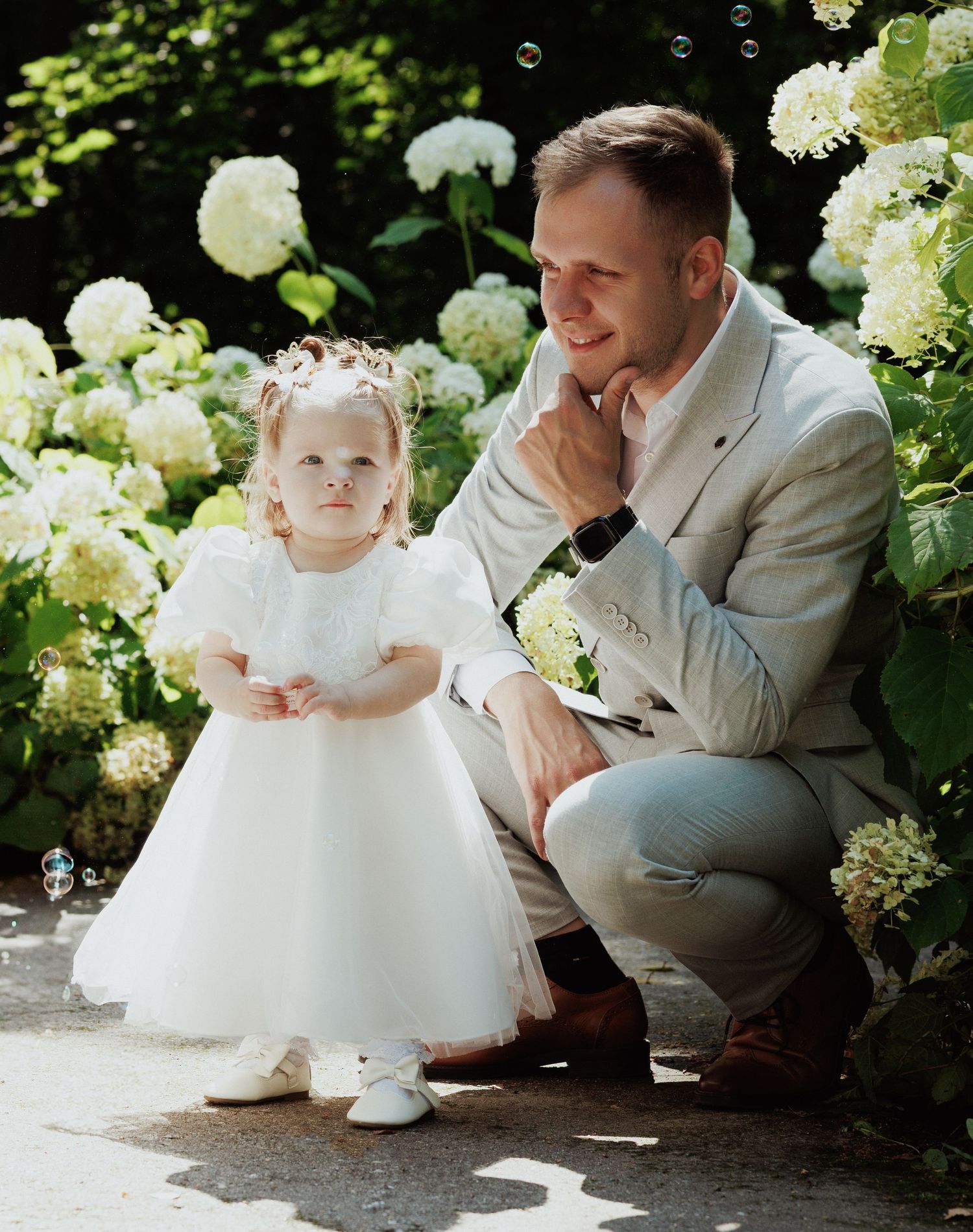 A tender moment in a garden with white hydrangea bushes as bubbles float through the sunlit air.