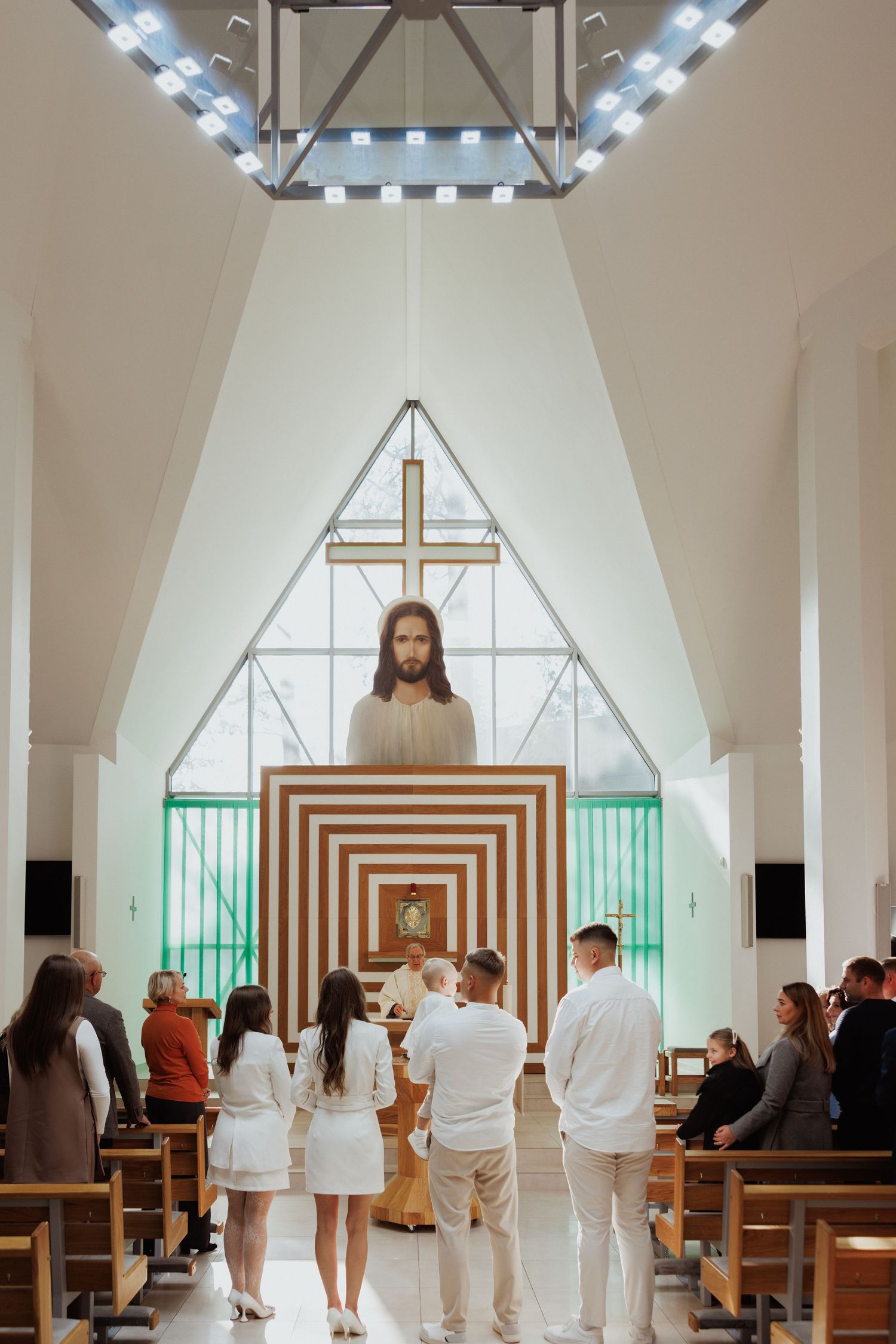 People stand in white attire inside a modern chapel with geometric wooden altar and skylights during a ceremony.