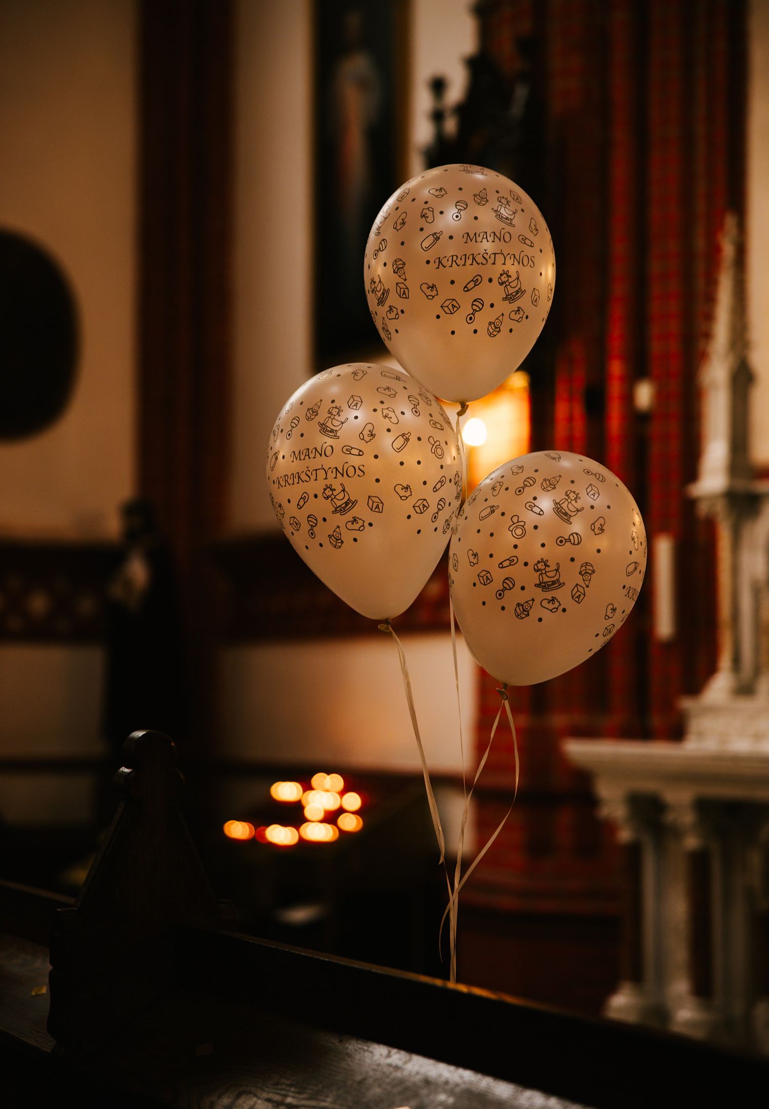 Three white balloons with text decorations float against a warm background with red curtains and glowing lights.