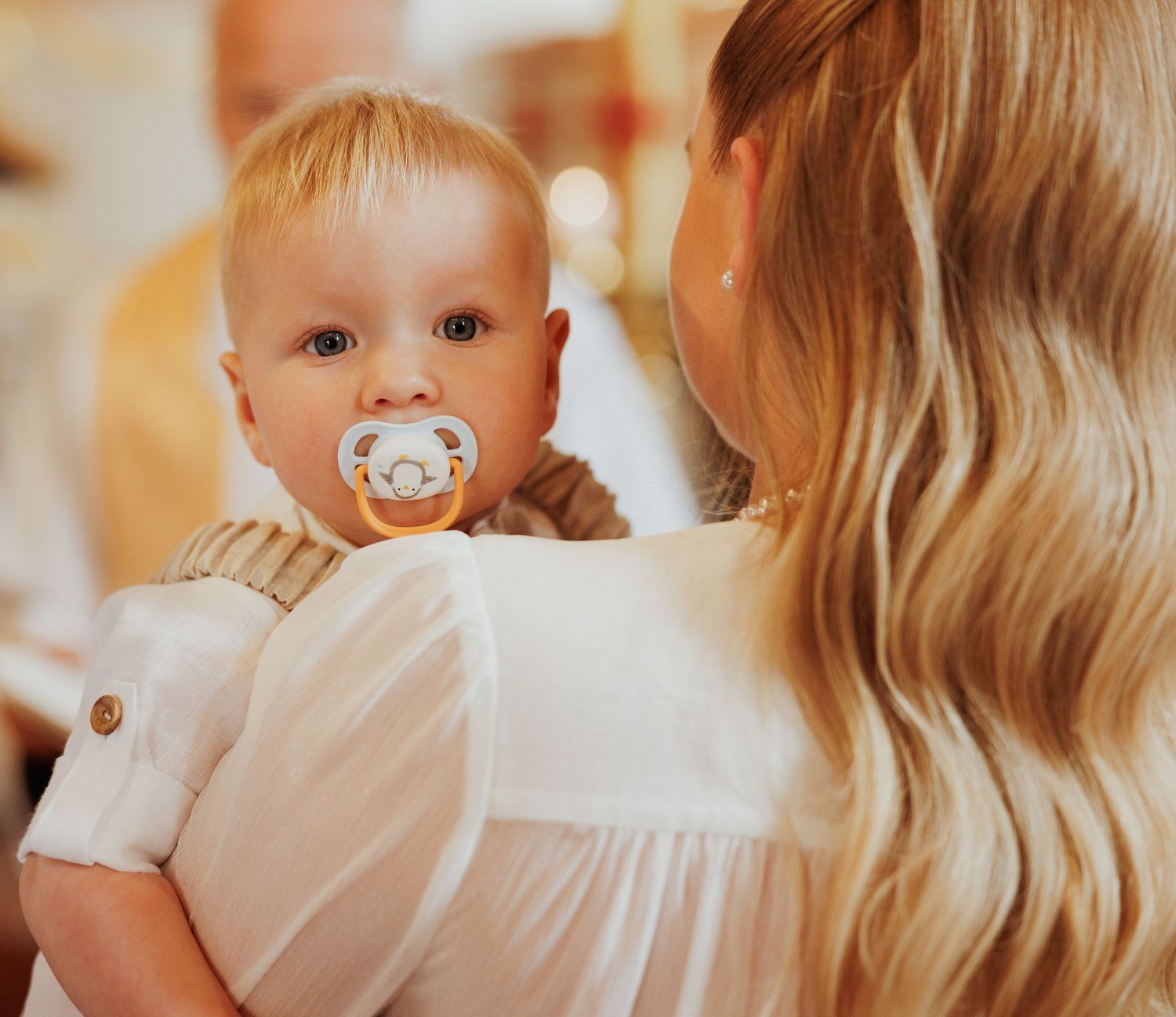A tender moment shows a baby with a pacifier being held in white clothing against a warm background.