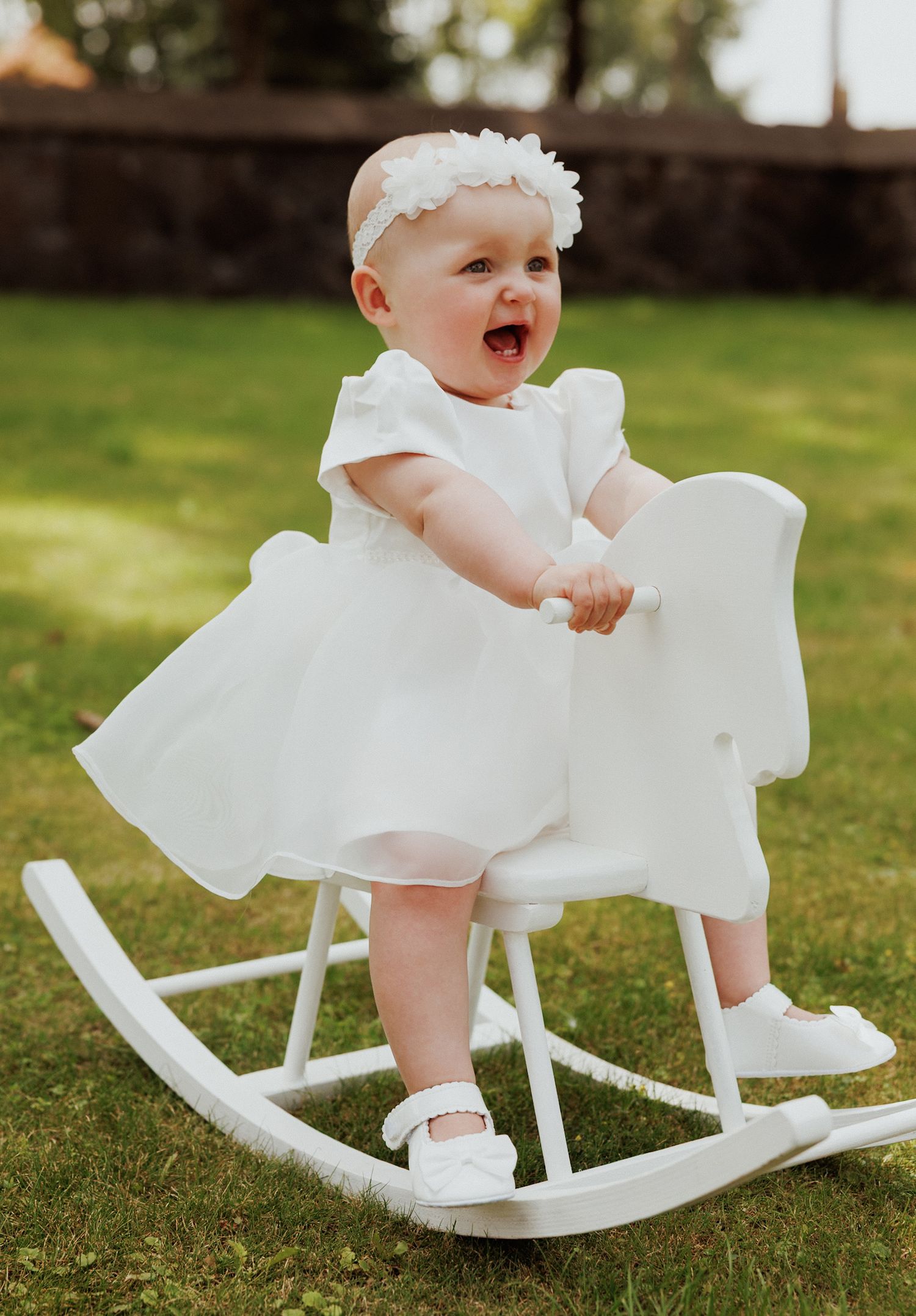 A baby in a white dress sits on a white wooden rocking horse outdoors on grass in multiple sequential photos.