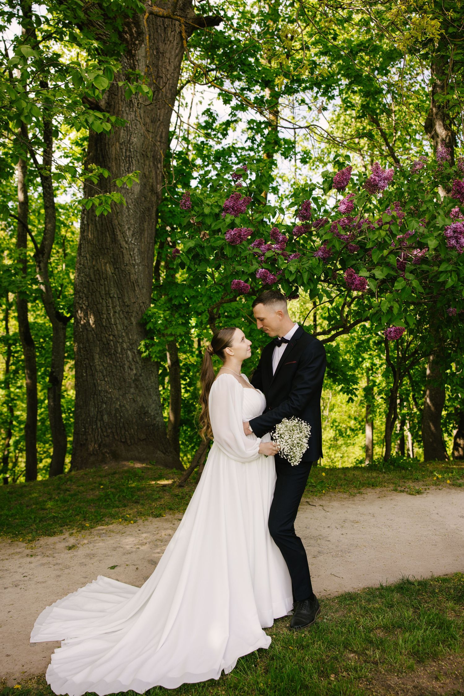 A wedding couple poses on a garden path with blooming trees and flowers in the background.