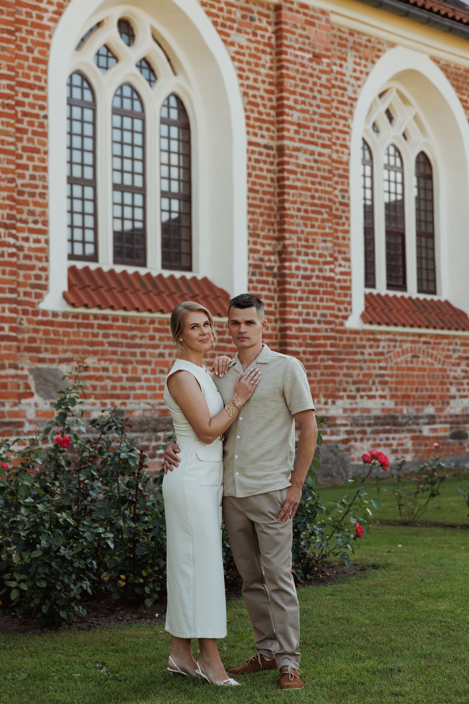 Couple poses together in front of a brick church with arched windows and rose bushes.