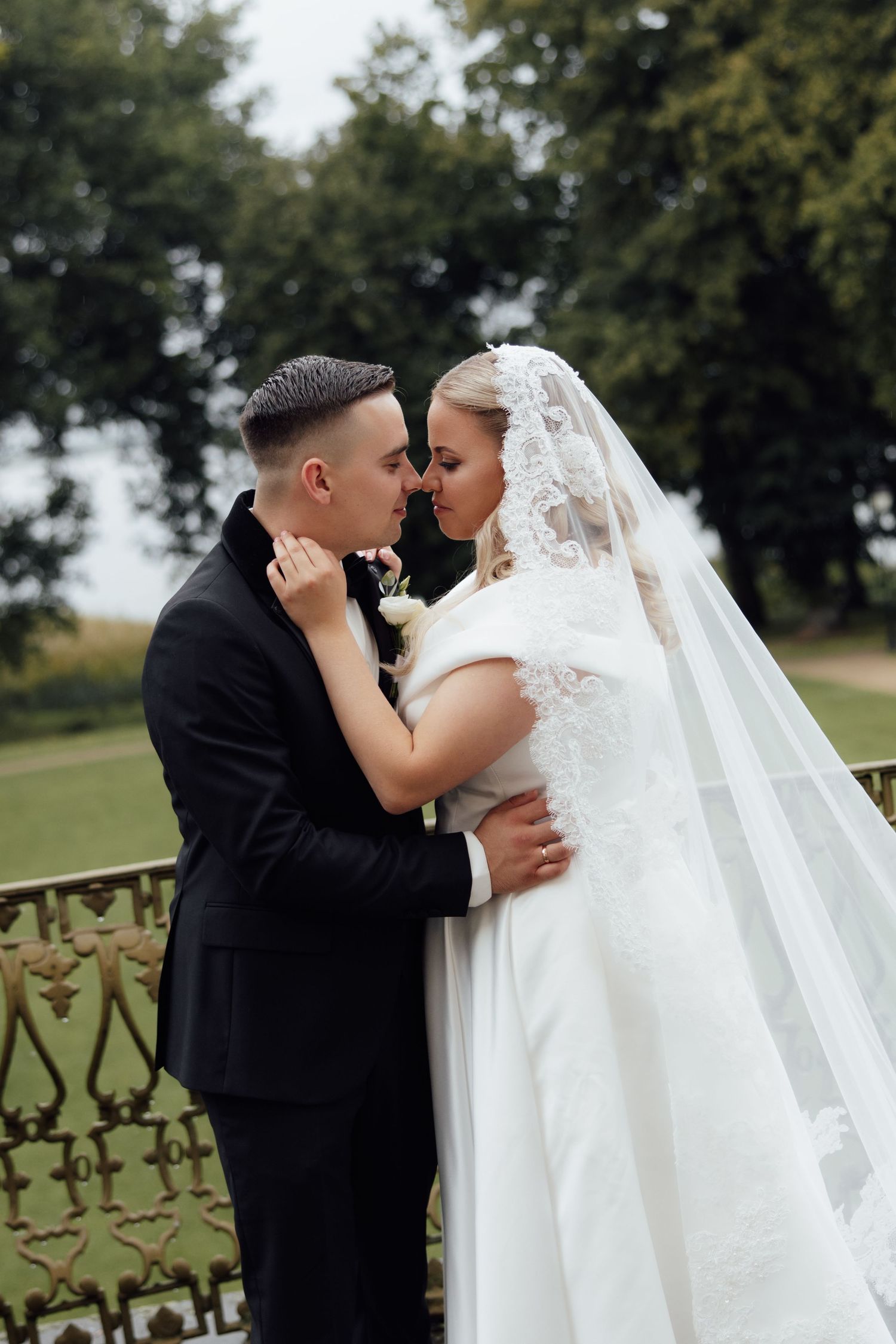 A bride in a white lace veil and groom in a black suit share a romantic moment on an ornate balcony outdoors.