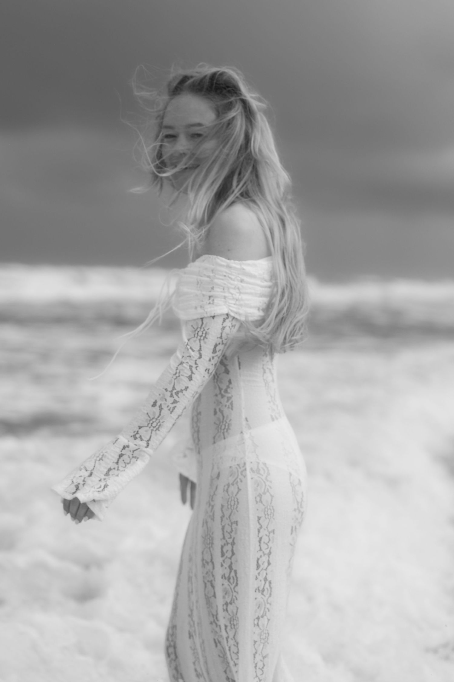 Black and white photo of a woman in a lace off-shoulder dress on a windy beach with ocean waves behind her.