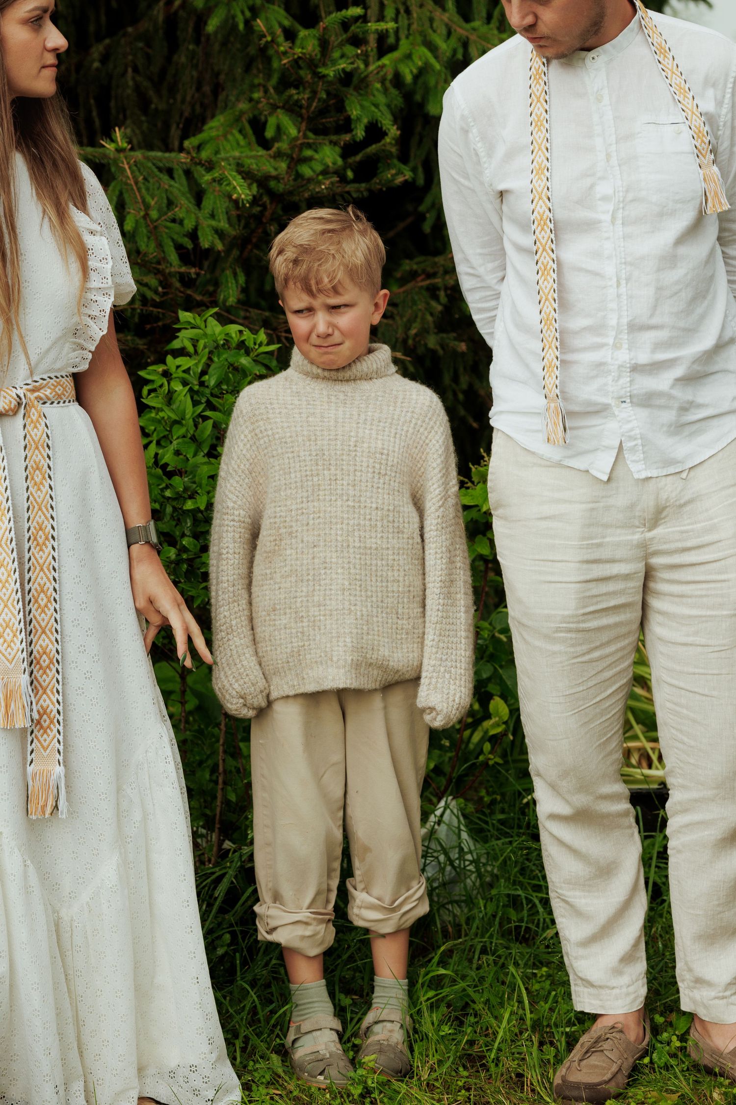 Young blonde boy in beige knit sweater standing between adults in white linen clothing outdoors.