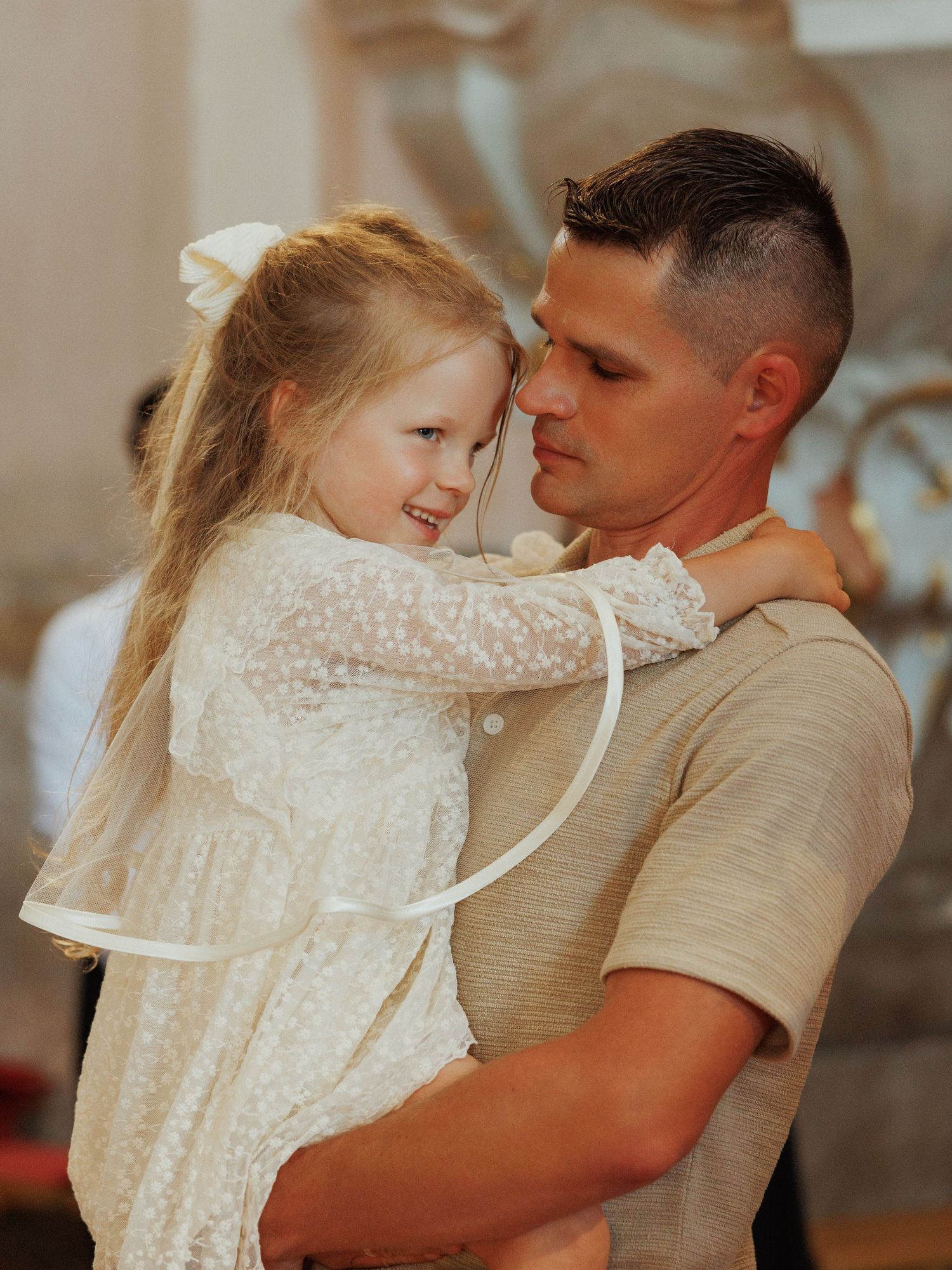 A tender moment captured between a parent and young child in a white lace dress embracing in warm lighting.