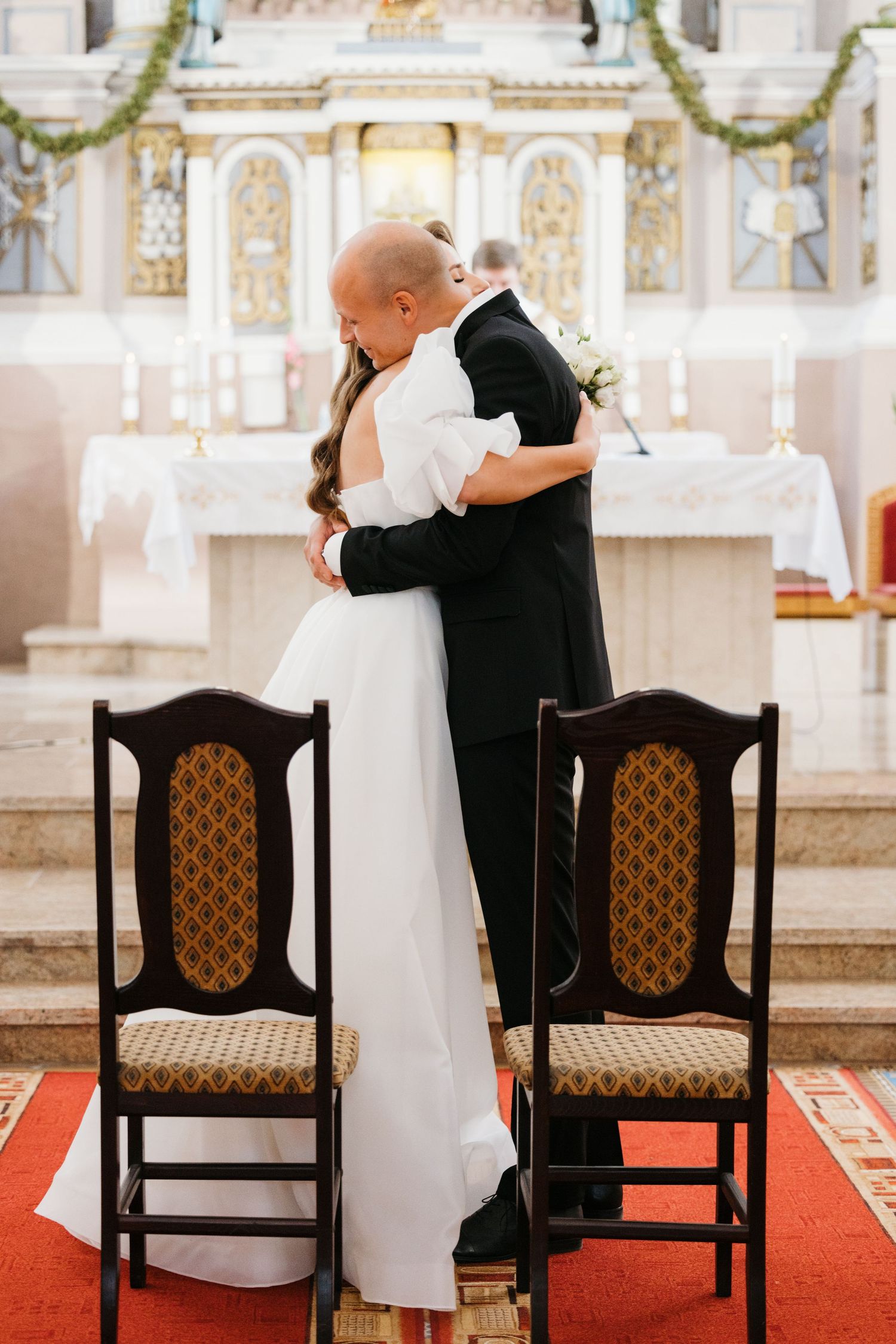 Intimate church ceremony moments captured with couple seated in traditional wooden chairs with ornate details.