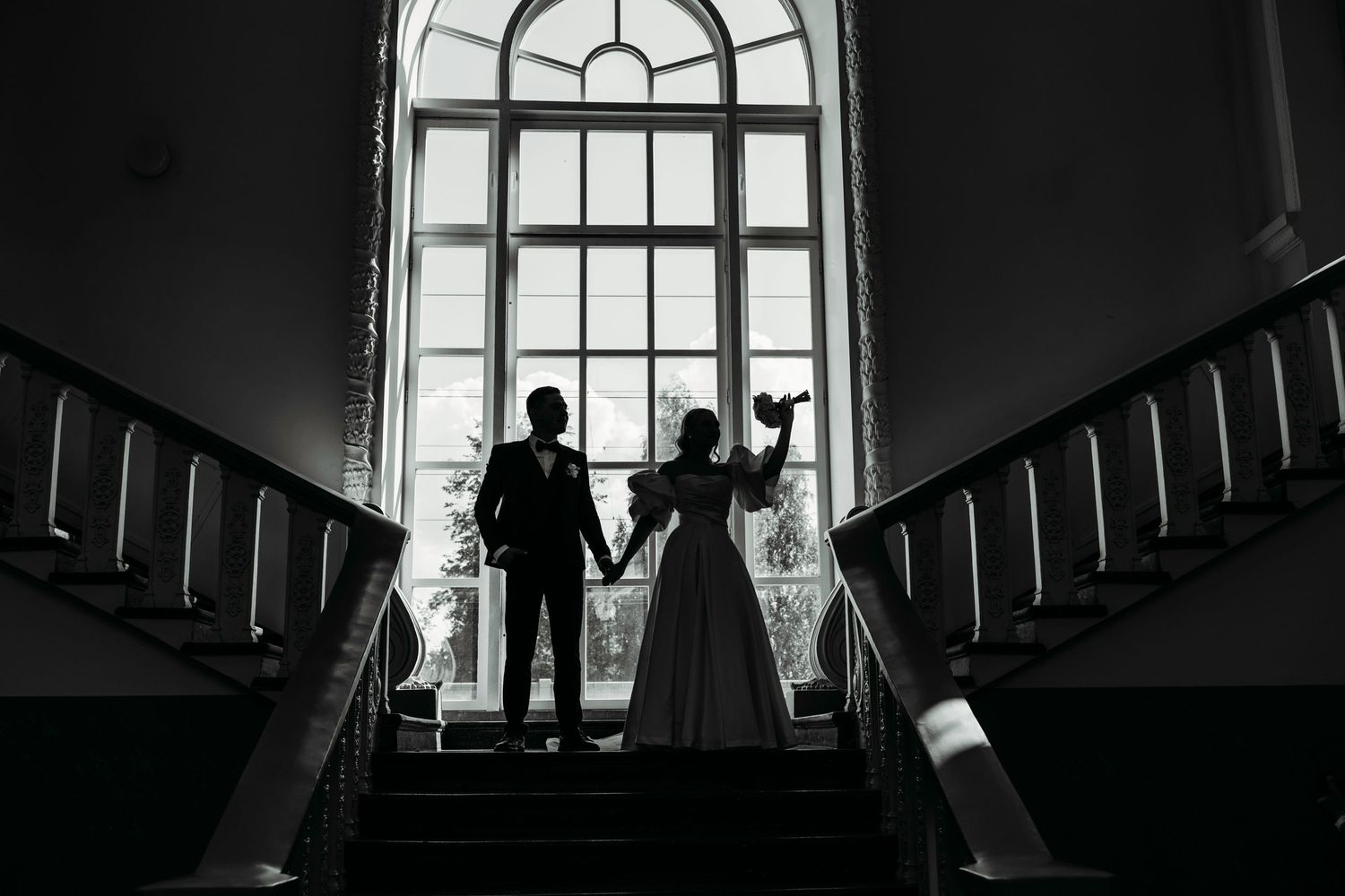 Silhouetted couple stands holding hands on grand staircase in front of arched window in black and white photo.