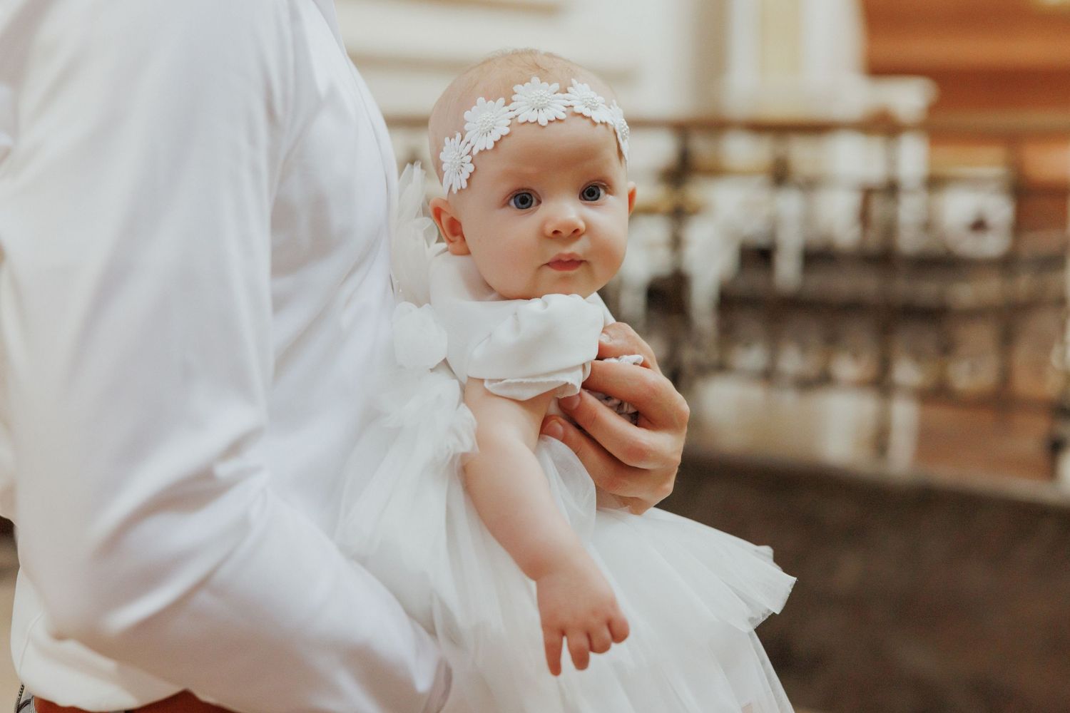 A small baby wearing a white christening gown sits on a wooden floor in warm lighting.