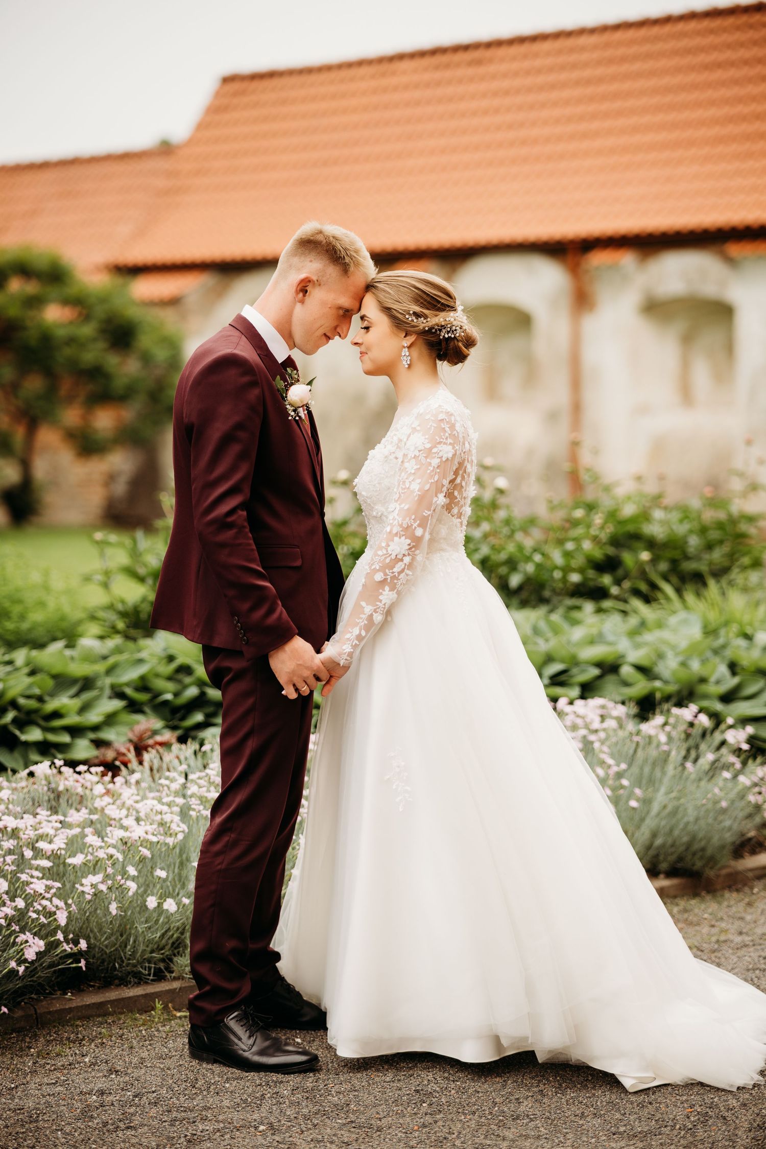 Wedding couple poses in garden with white daisies and brick building backdrop.