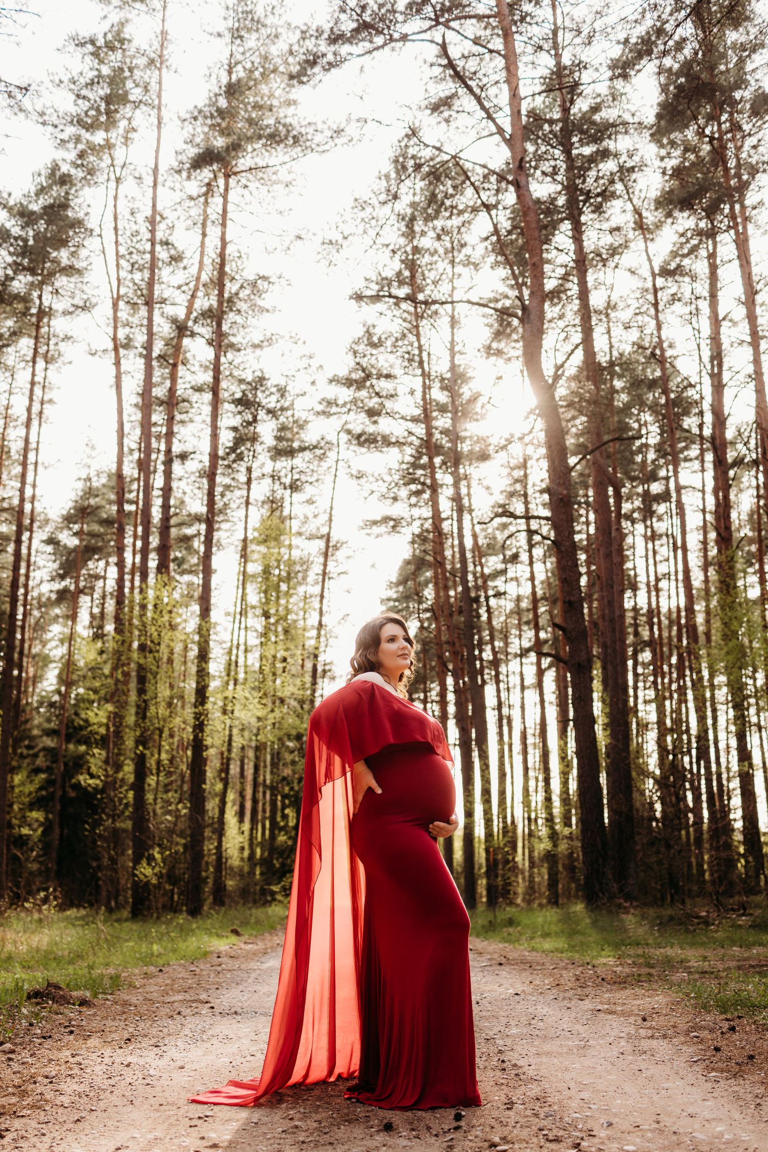 Dramatic maternity photos in a red dress along a forest path with tall pine trees.